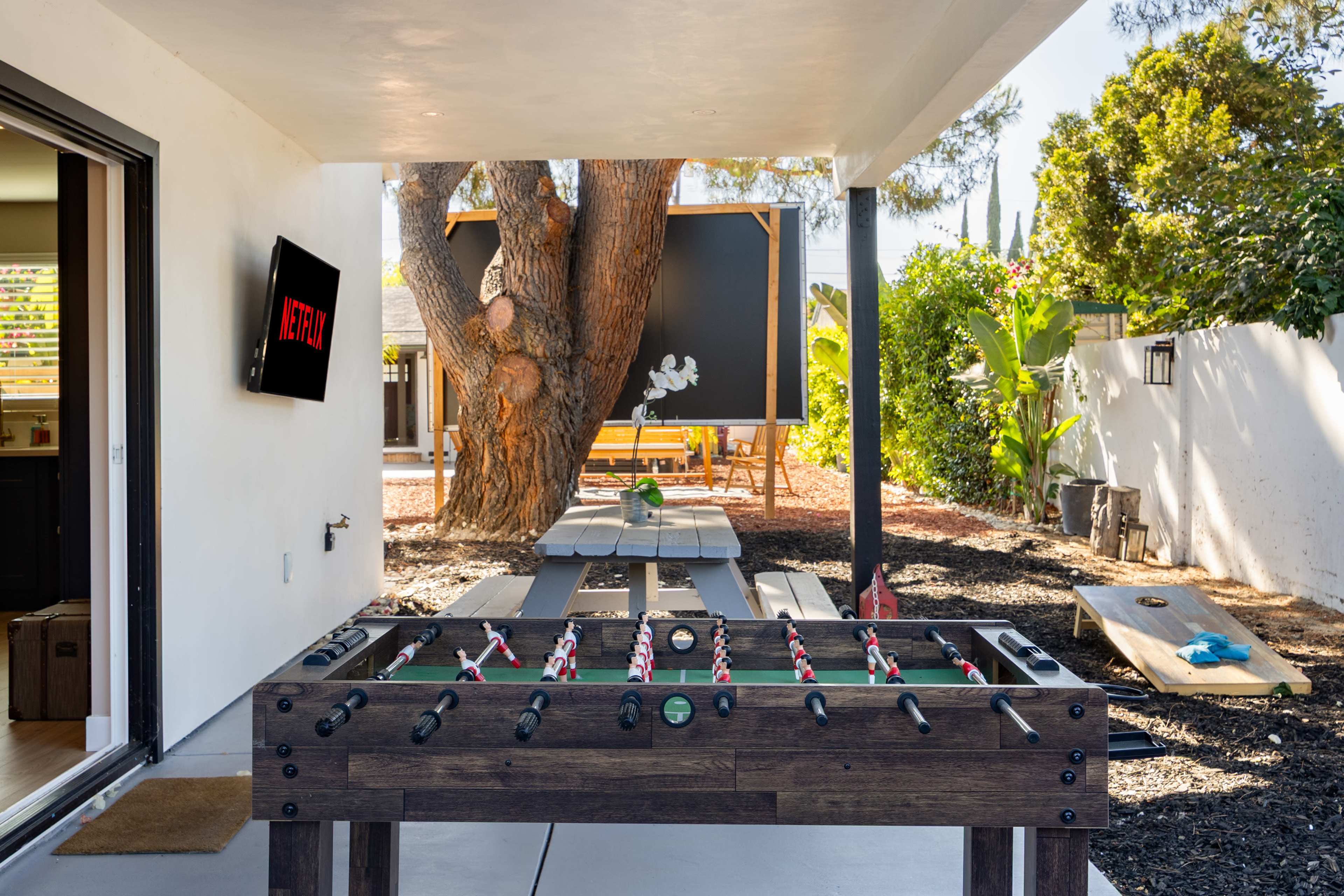 A foosball table is positioned under an outdoor patio, with a large tree and picnic table visible in the background.