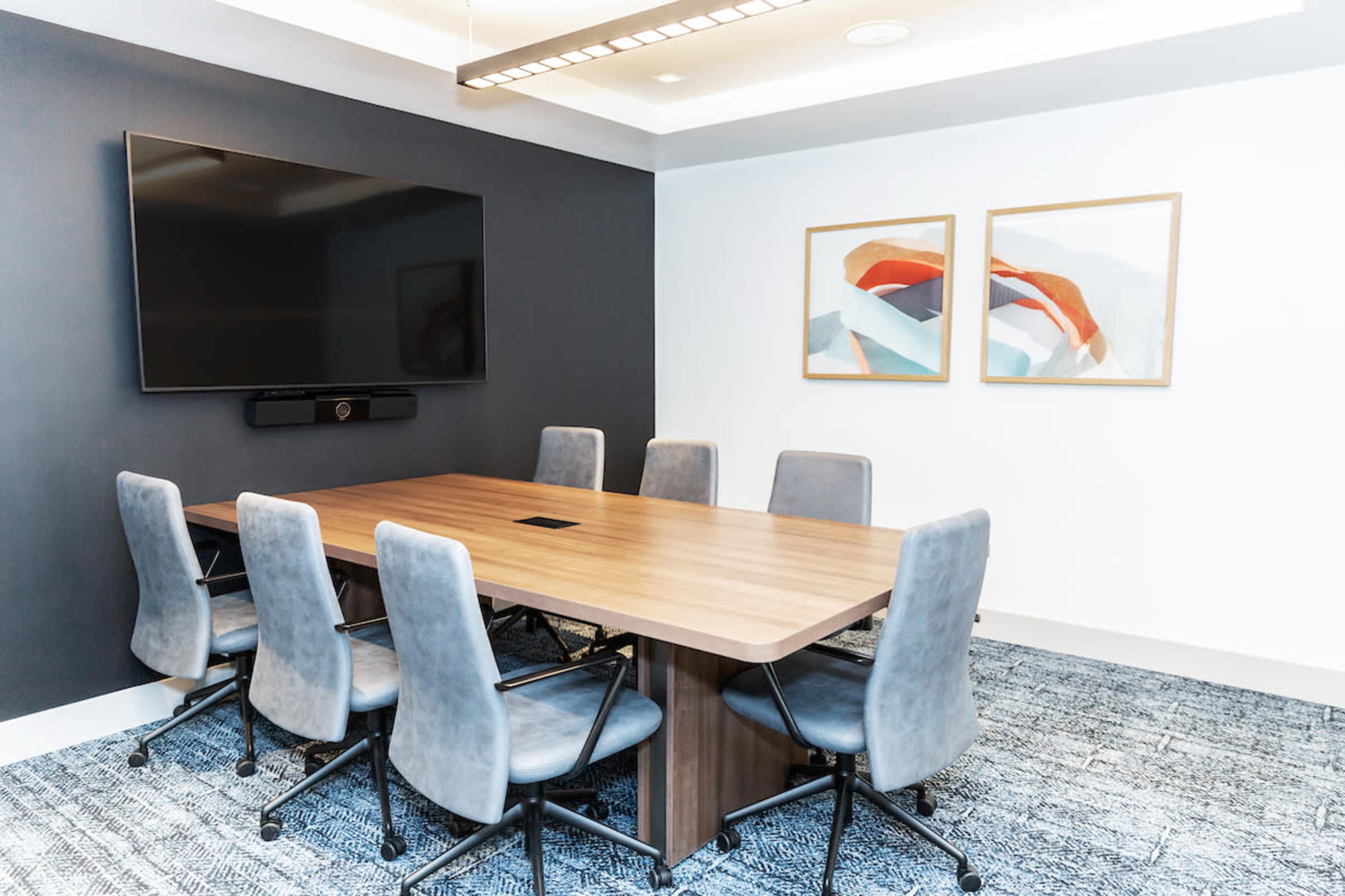 A modern conference room with a large wooden table surrounded by six gray chairs, a wall-mounted television, and two abstract paintings.
