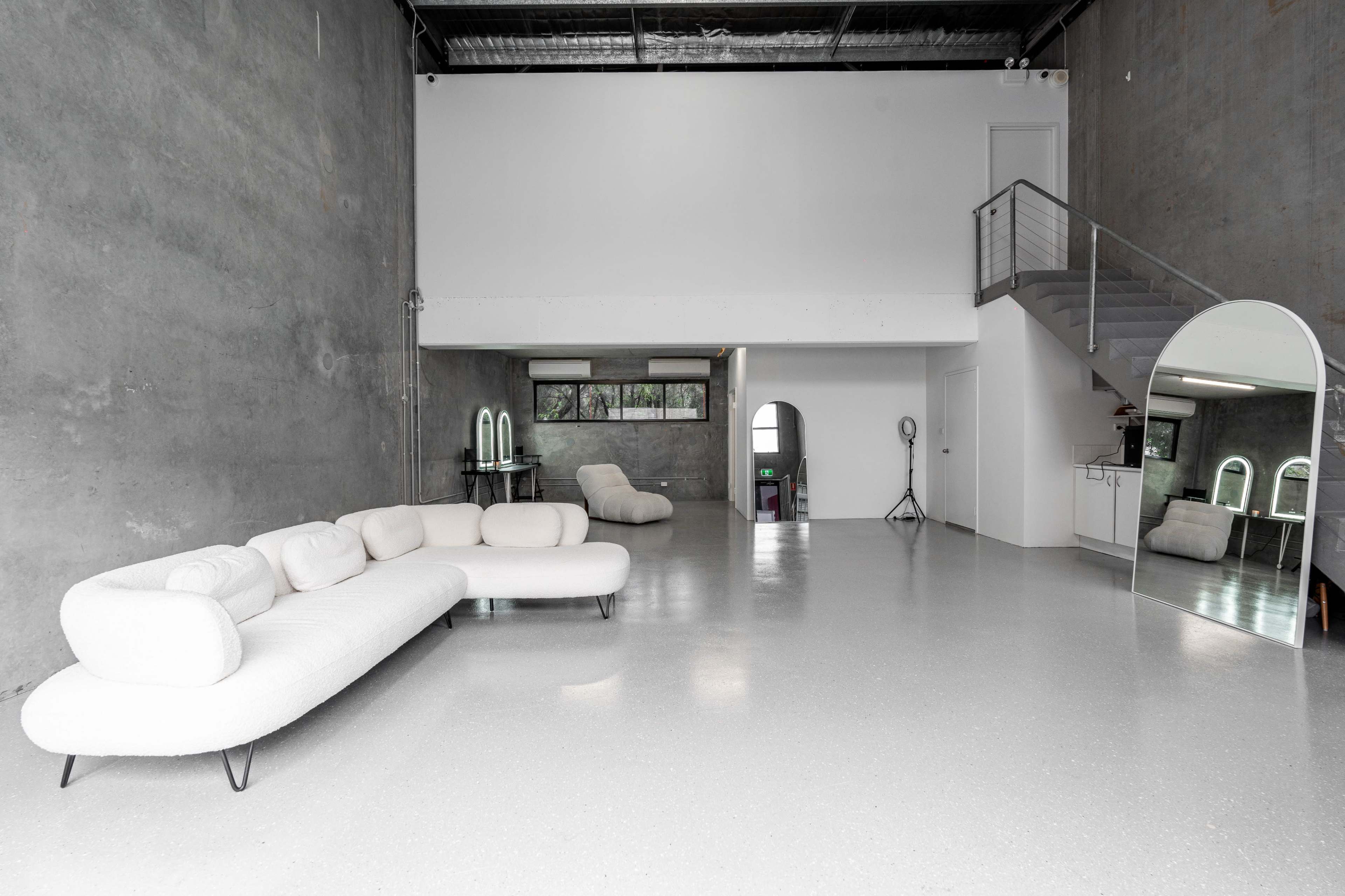 A spacious, minimalist interior with a light-colored sectional sofa, a staircase, and a large mirror against a textured gray wall.