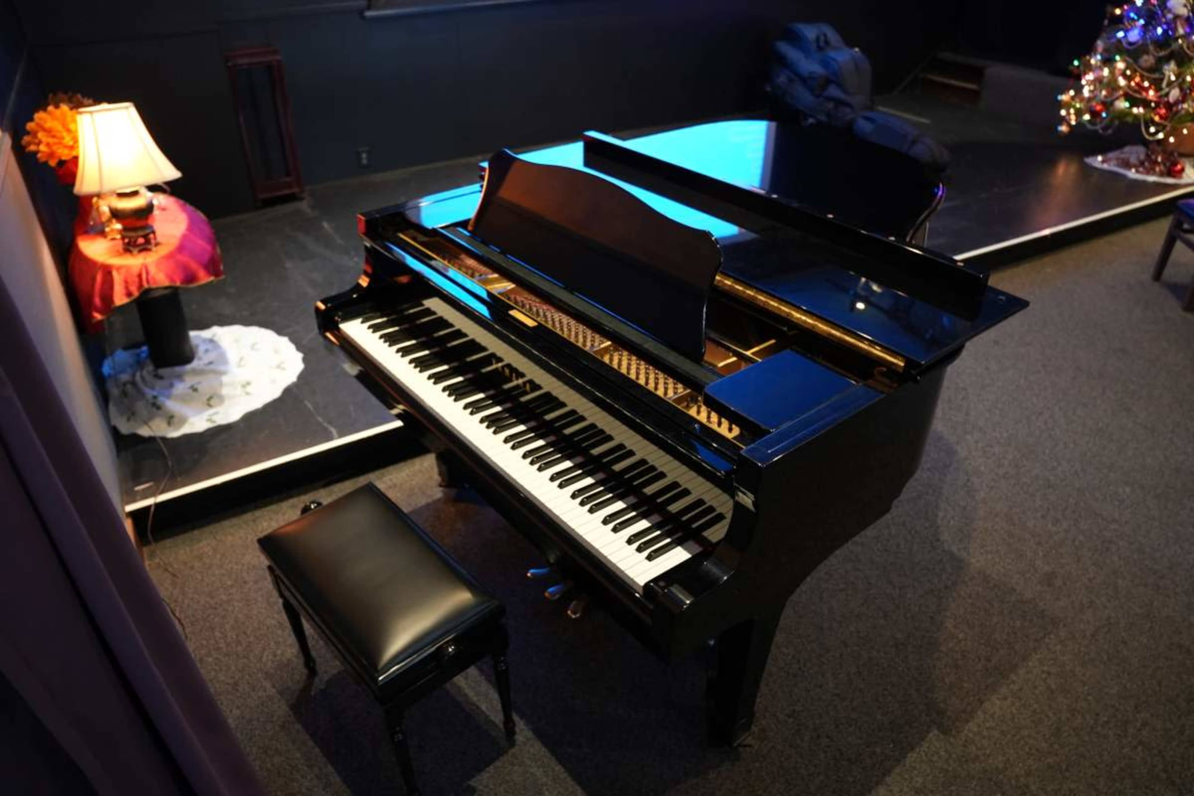 A black grand piano with a matching bench is situated on a carpeted stage, with a lamp and a decorated Christmas tree in the background.