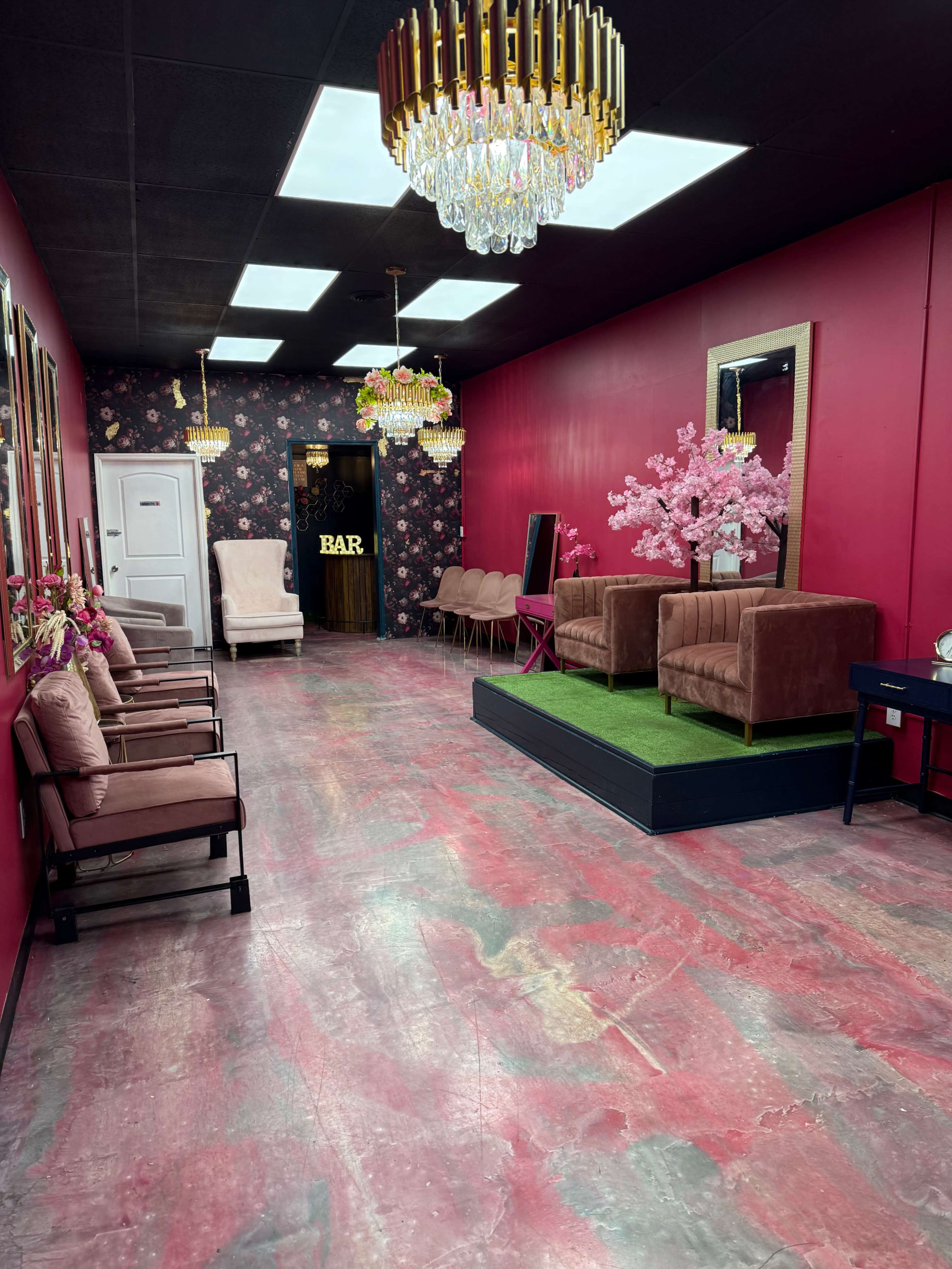 The image shows a brightly colored salon with pink walls, multiple chandeliers, and two leather chairs separated by a faux grass area.