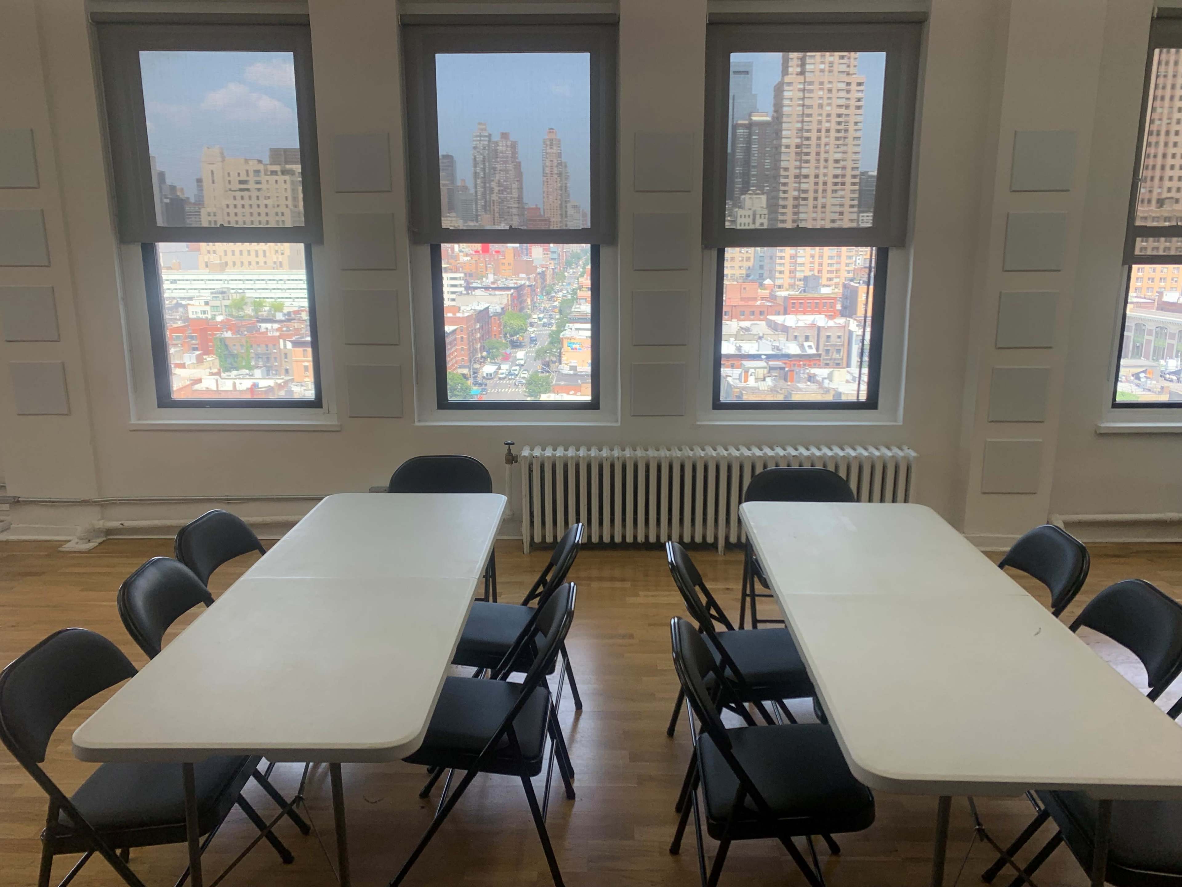 A room with two empty tables and chairs arranged in the center, and large windows providing a view of a city skyline outside.