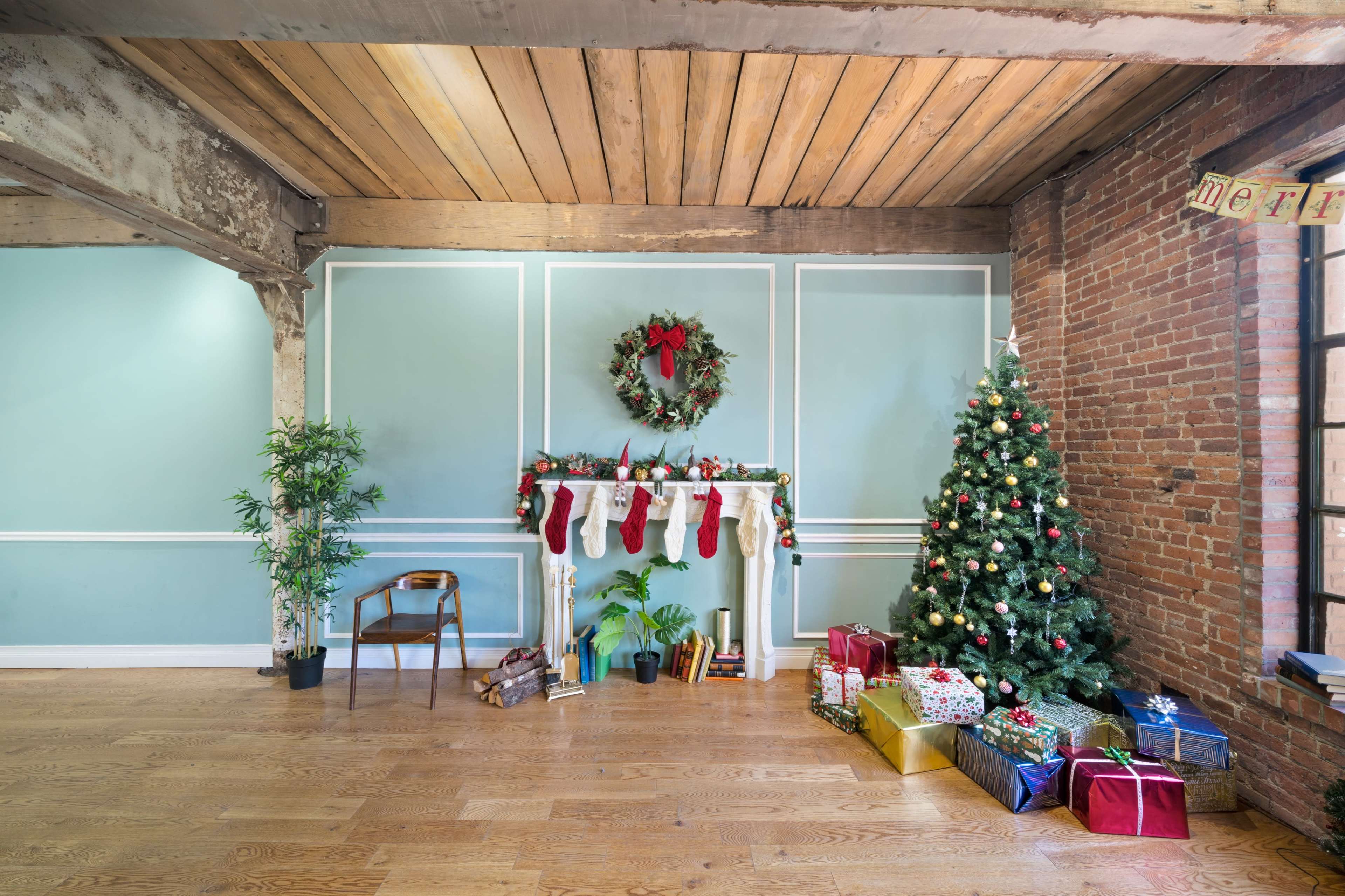 A decorated holiday scene features a Christmas tree, a mantel with stockings, and wrapped gifts in a spacious room with wooden floors and exposed brick walls.