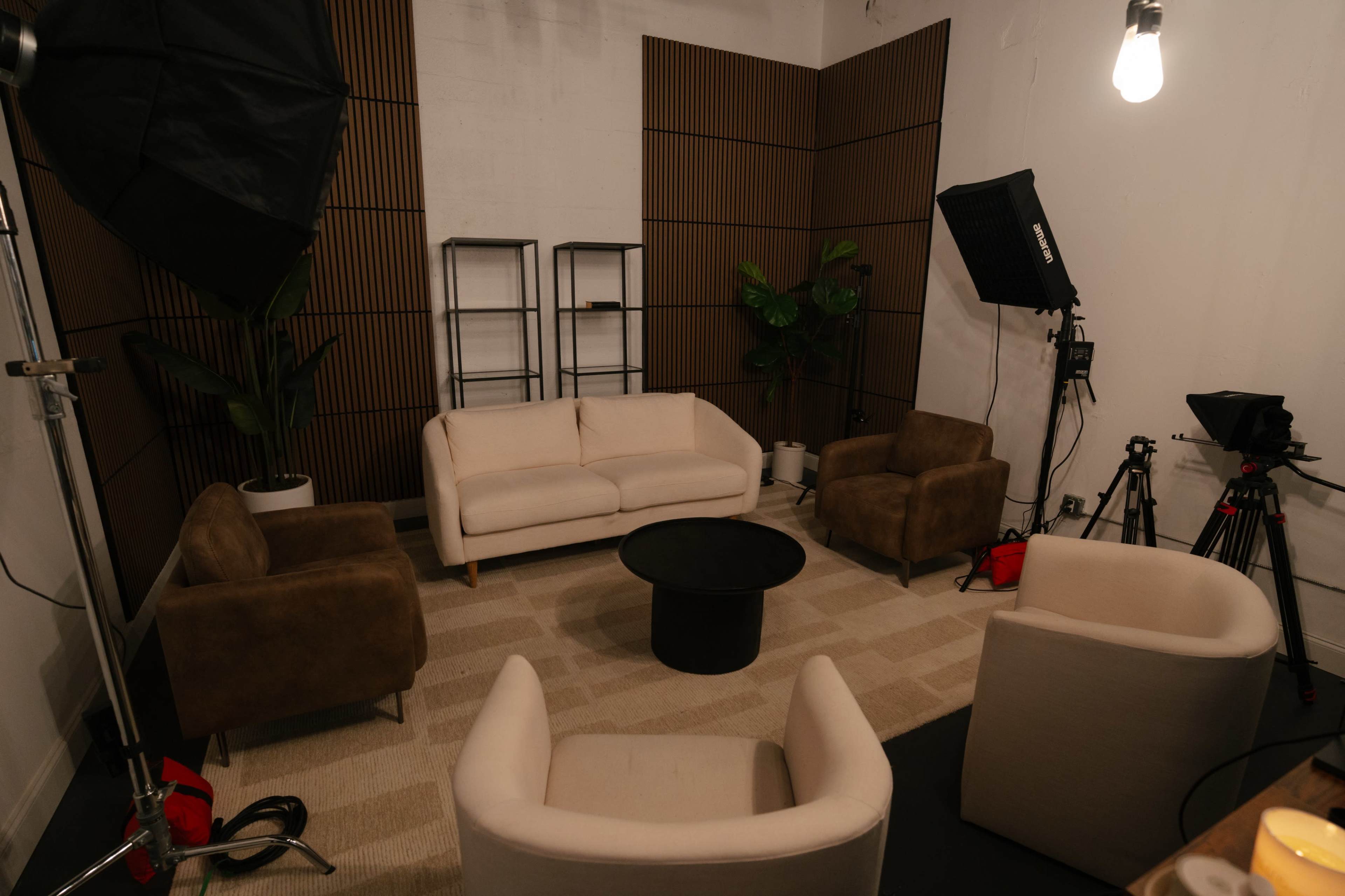 The image shows a modern studio setup with a beige sectional sofa, three brown armchairs, a black round coffee table, and various lighting equipment in a minimally decorated space.