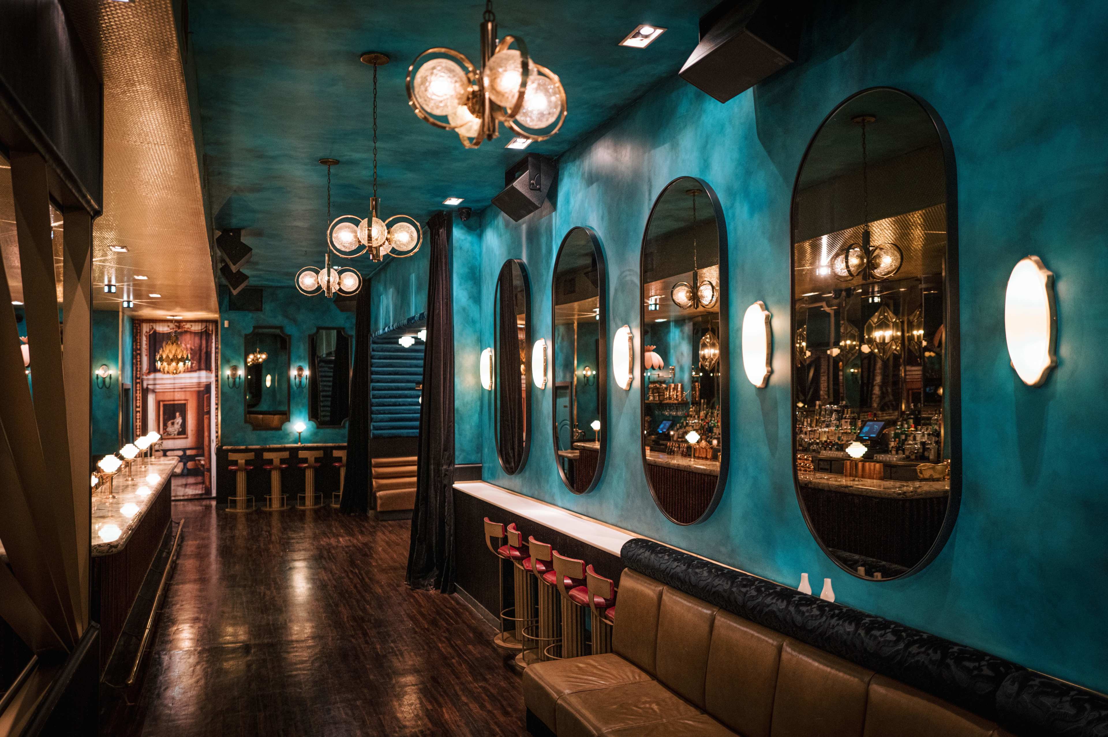 The image depicts a stylish bar interior featuring turquoise walls, multiple oval mirrors, and a long seating area with stools.
