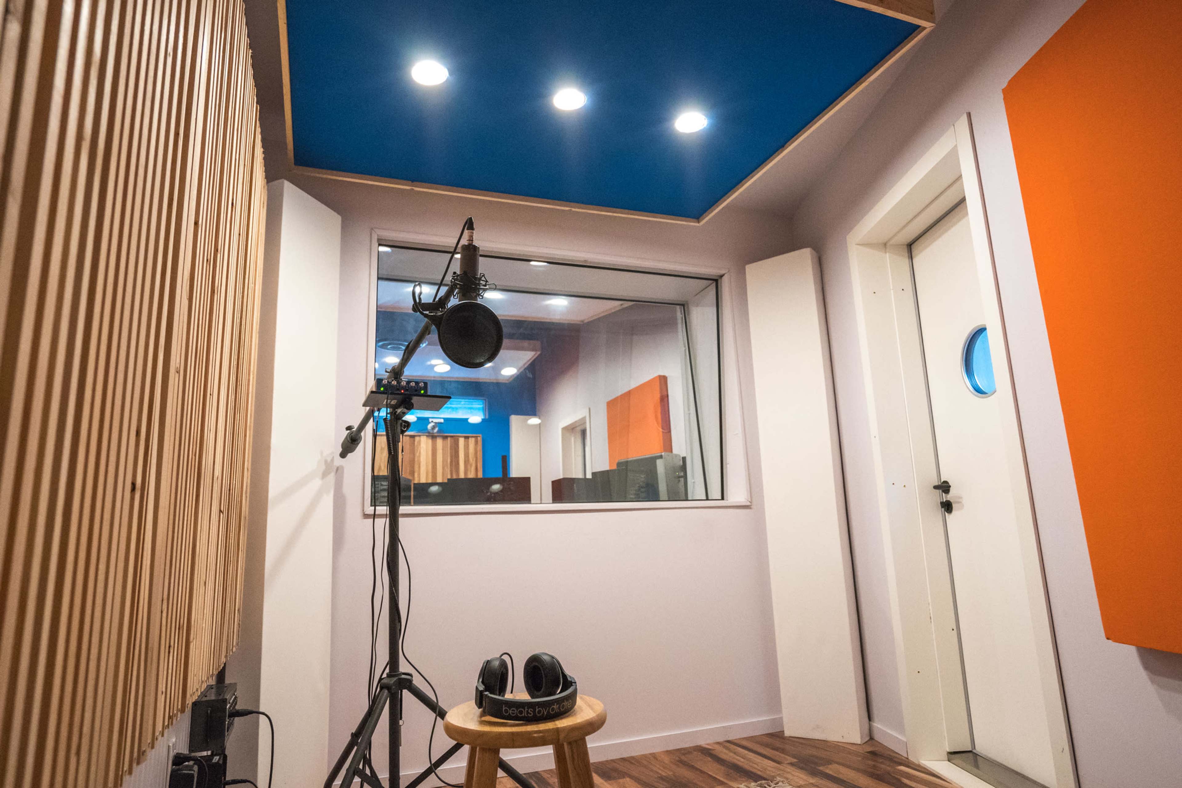 A small sound recording booth with a microphone on a stand, headphones on a stool, and a large window overlooking a control room.