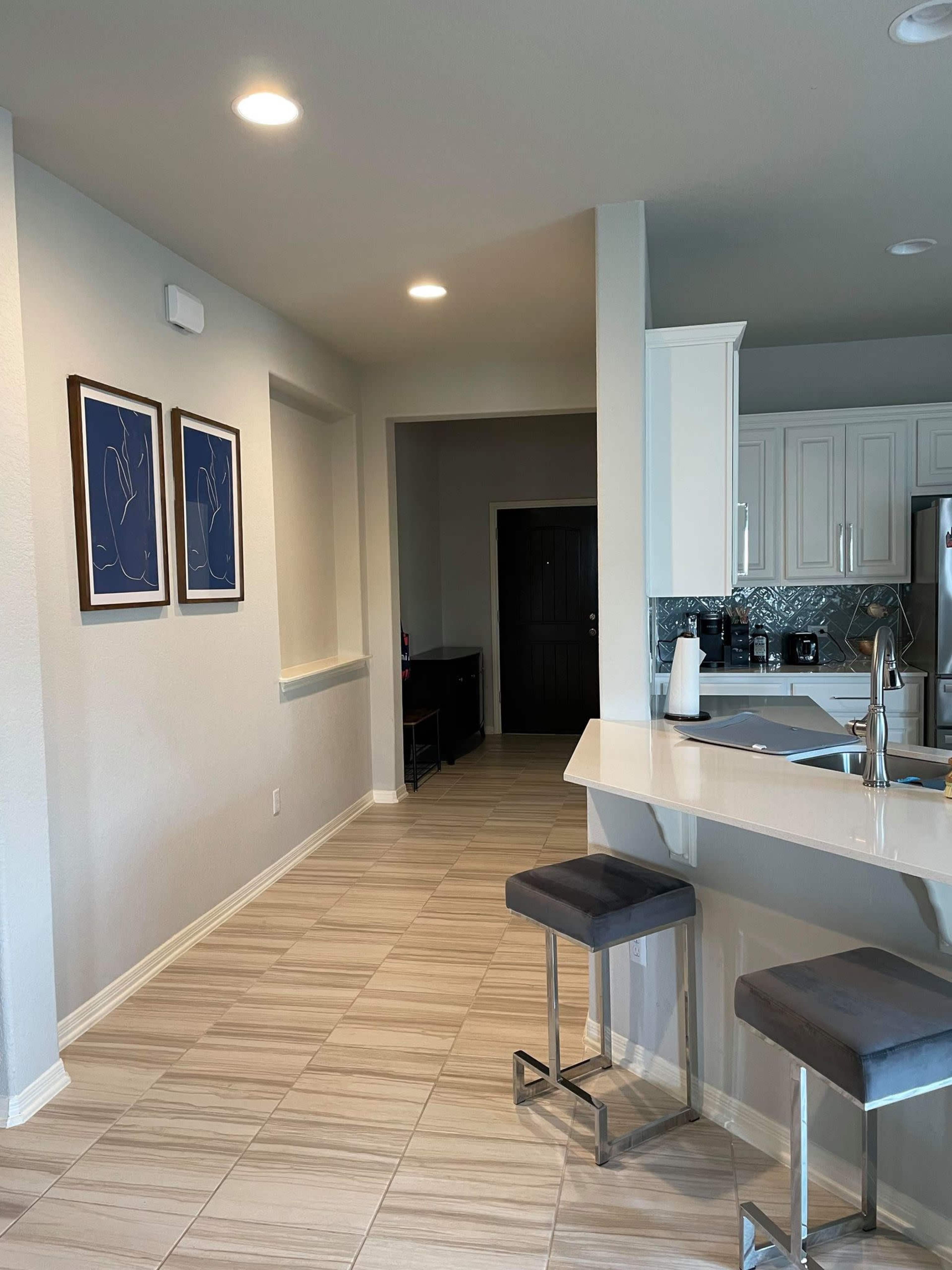 The image shows a modern kitchen with white cabinets, a bar with two gray stools, and a light-colored tiled floor leading to a hallway.