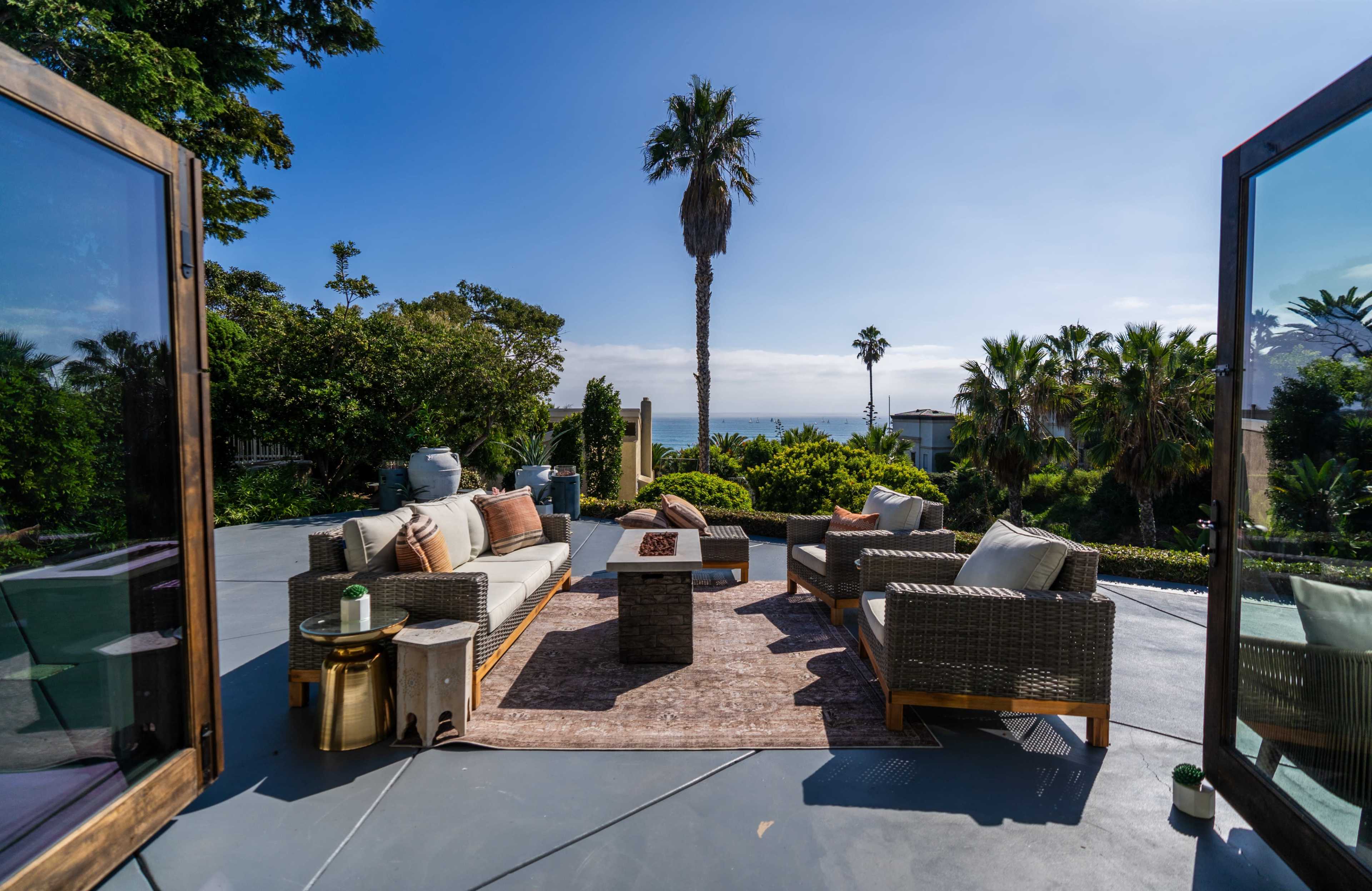 A patio with comfortable seating and a coffee table overlooks a blue ocean and palm trees under a clear sky.