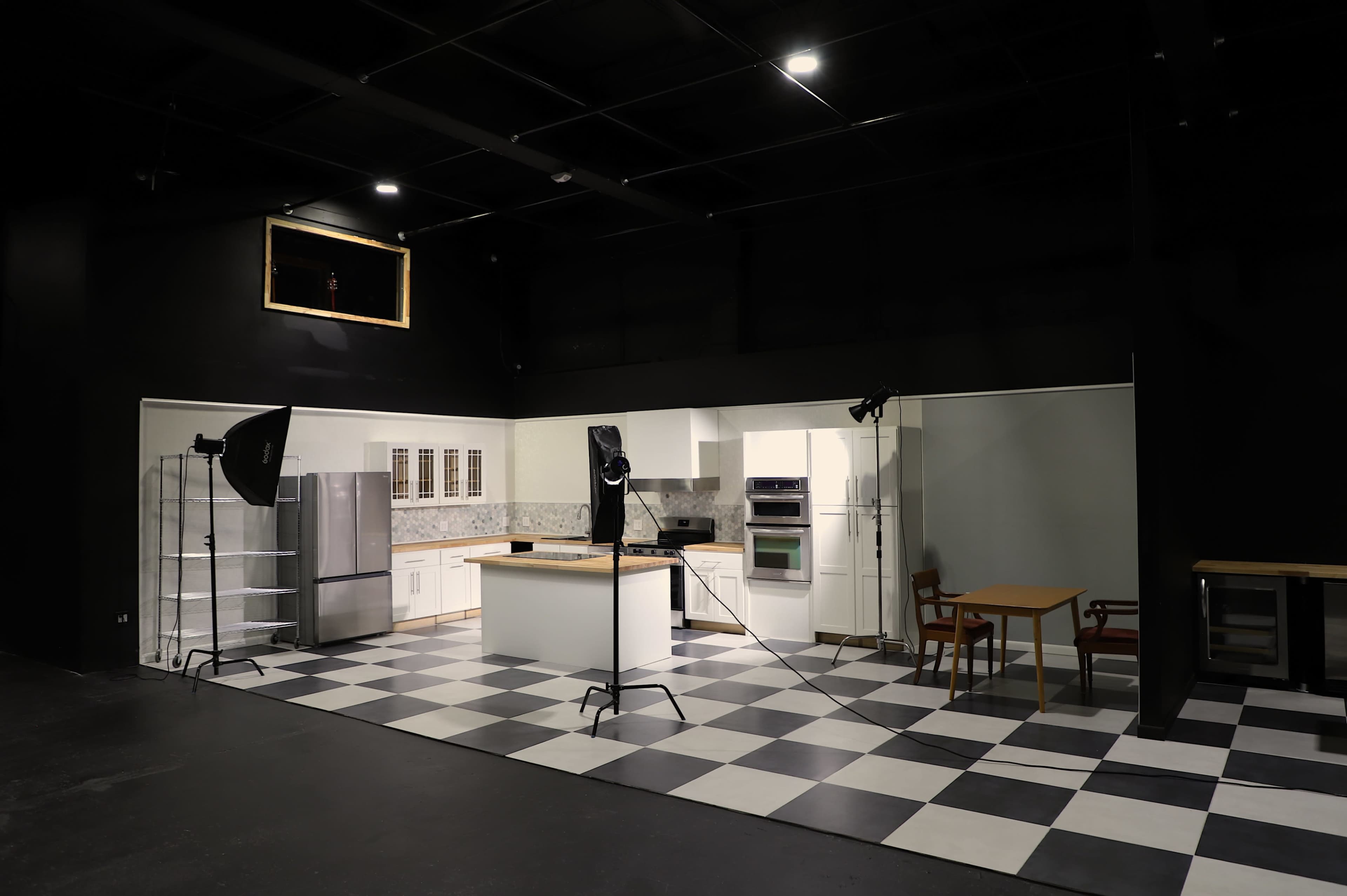 The image shows a modern kitchen set designed for a photo shoot, featuring a checkerboard floor, an island in the center, and professional lighting equipment positioned around the space.