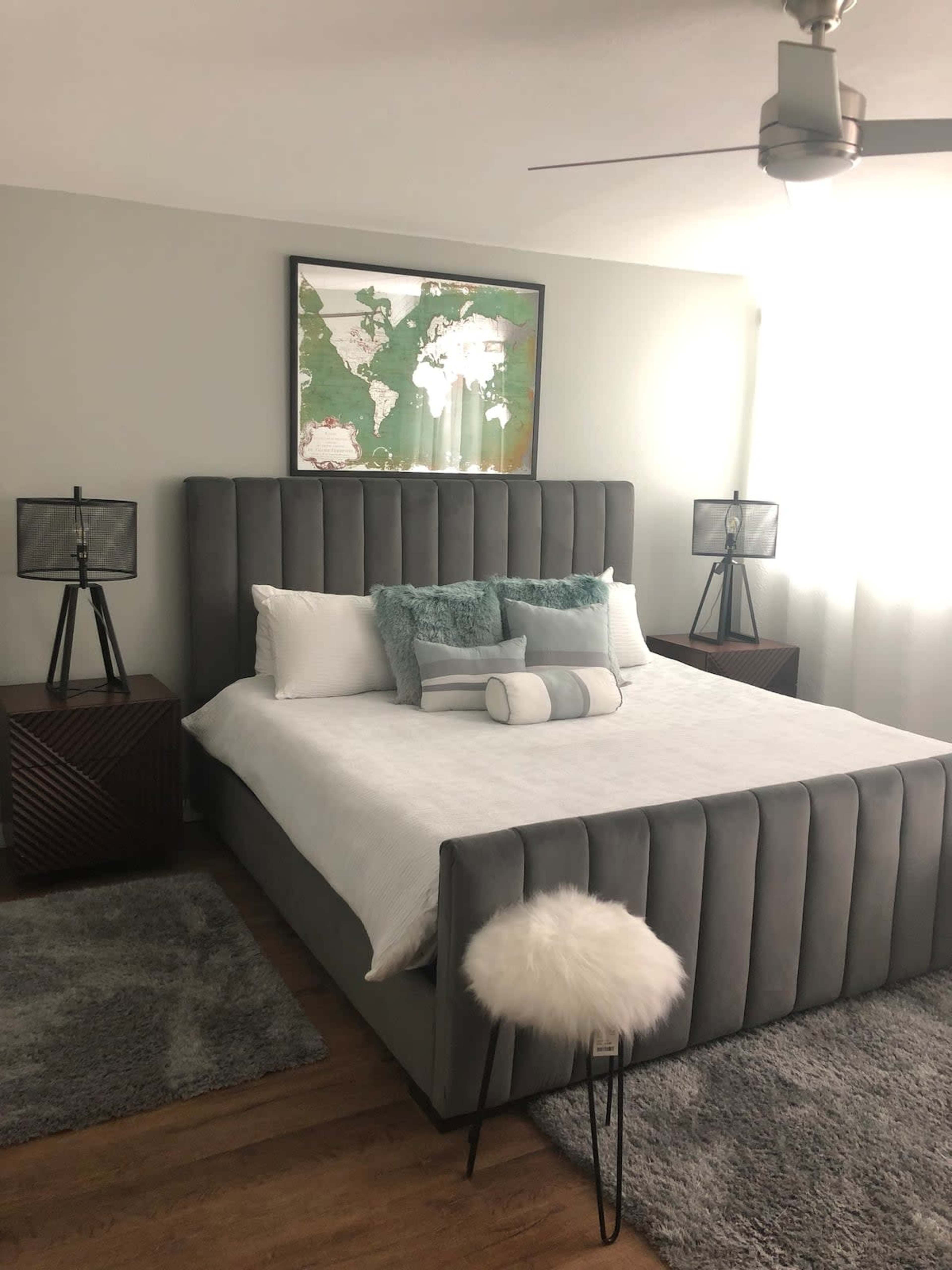 A modern bedroom featuring a gray upholstered bed, two bedside lamps, a map on the wall, and a fluffy stool near a wooden nightstand.