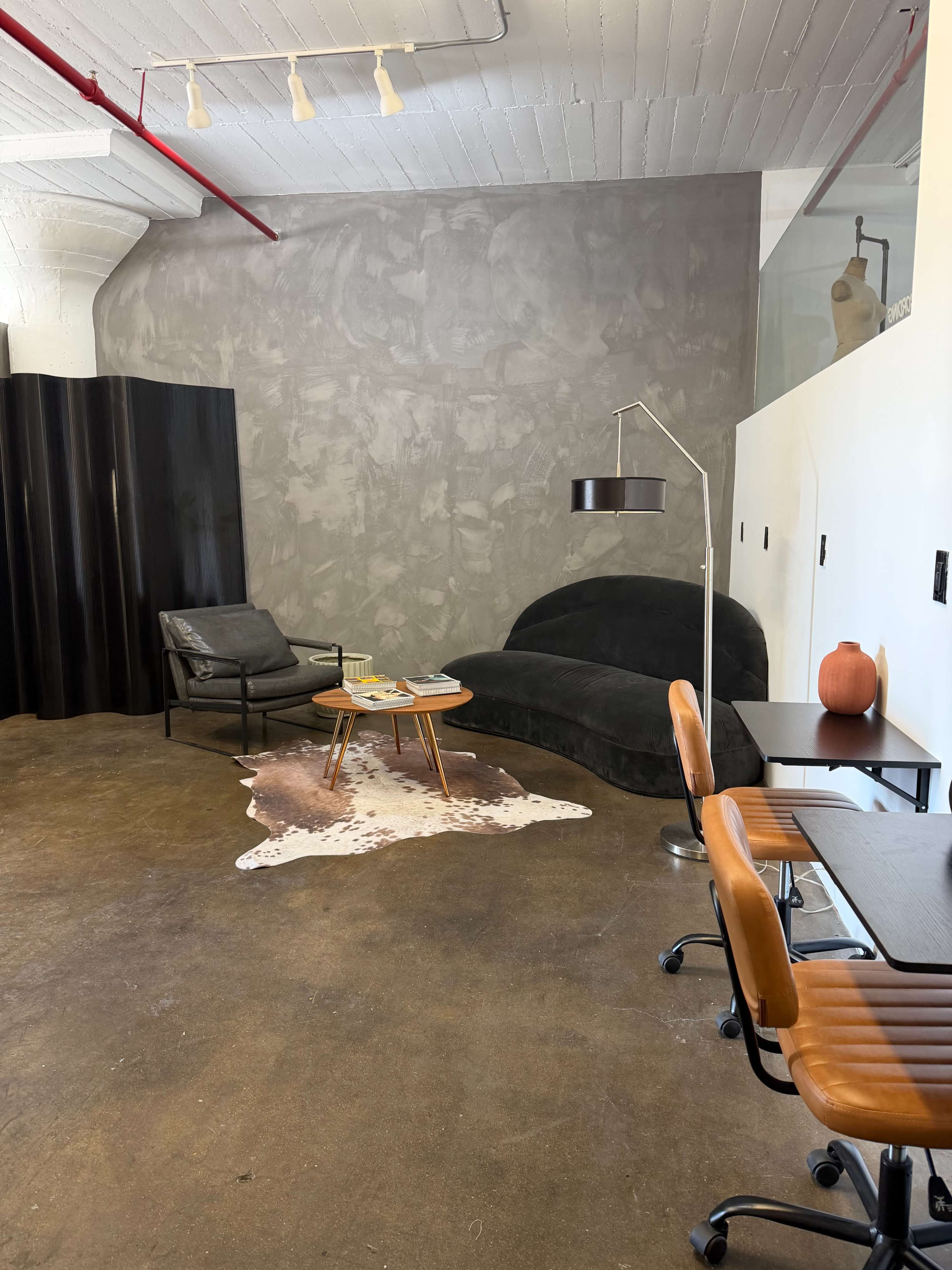 The image shows a minimalist interior space featuring a black sofa, a leather chair, a coffee table with magazines, and a cowhide rug on a concrete floor.