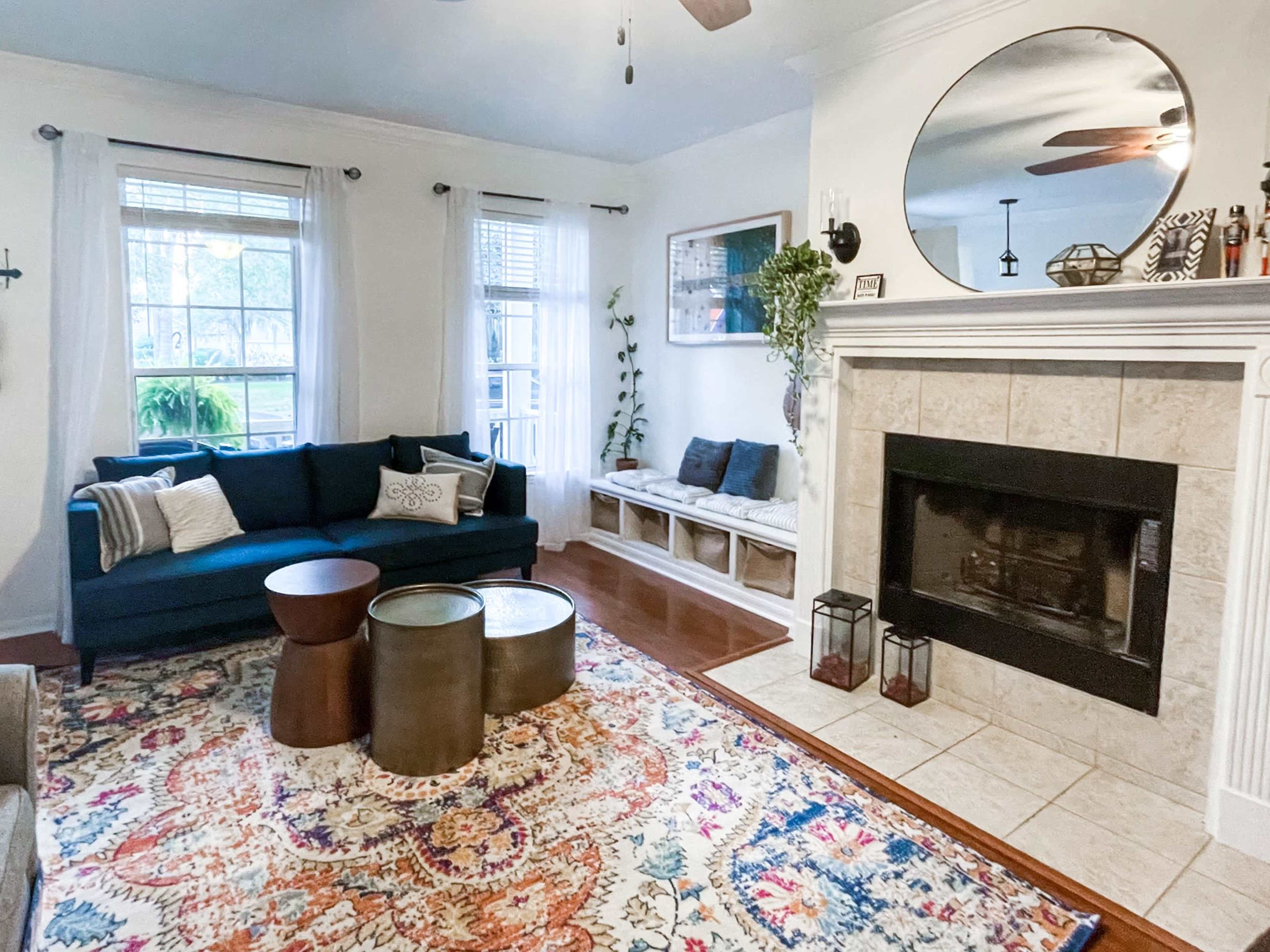 The image shows a living room with a teal sofa, two round metallic coffee tables, a large mirror above a tiled fireplace, and a colorful area rug.