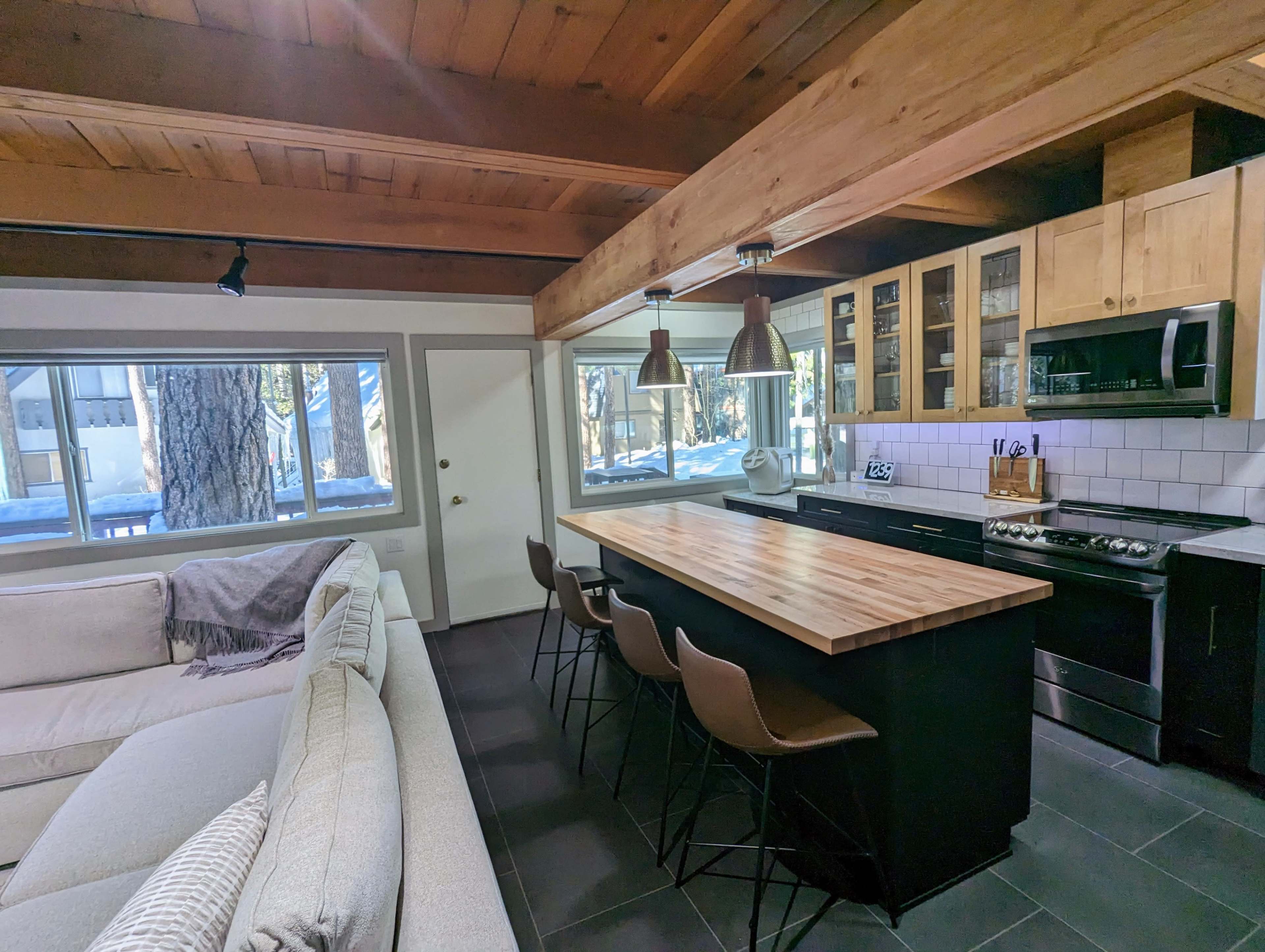 A modern kitchen with a wooden island, bar stools, and stainless steel appliances, situated beside a cozy living area featuring a beige sofa.