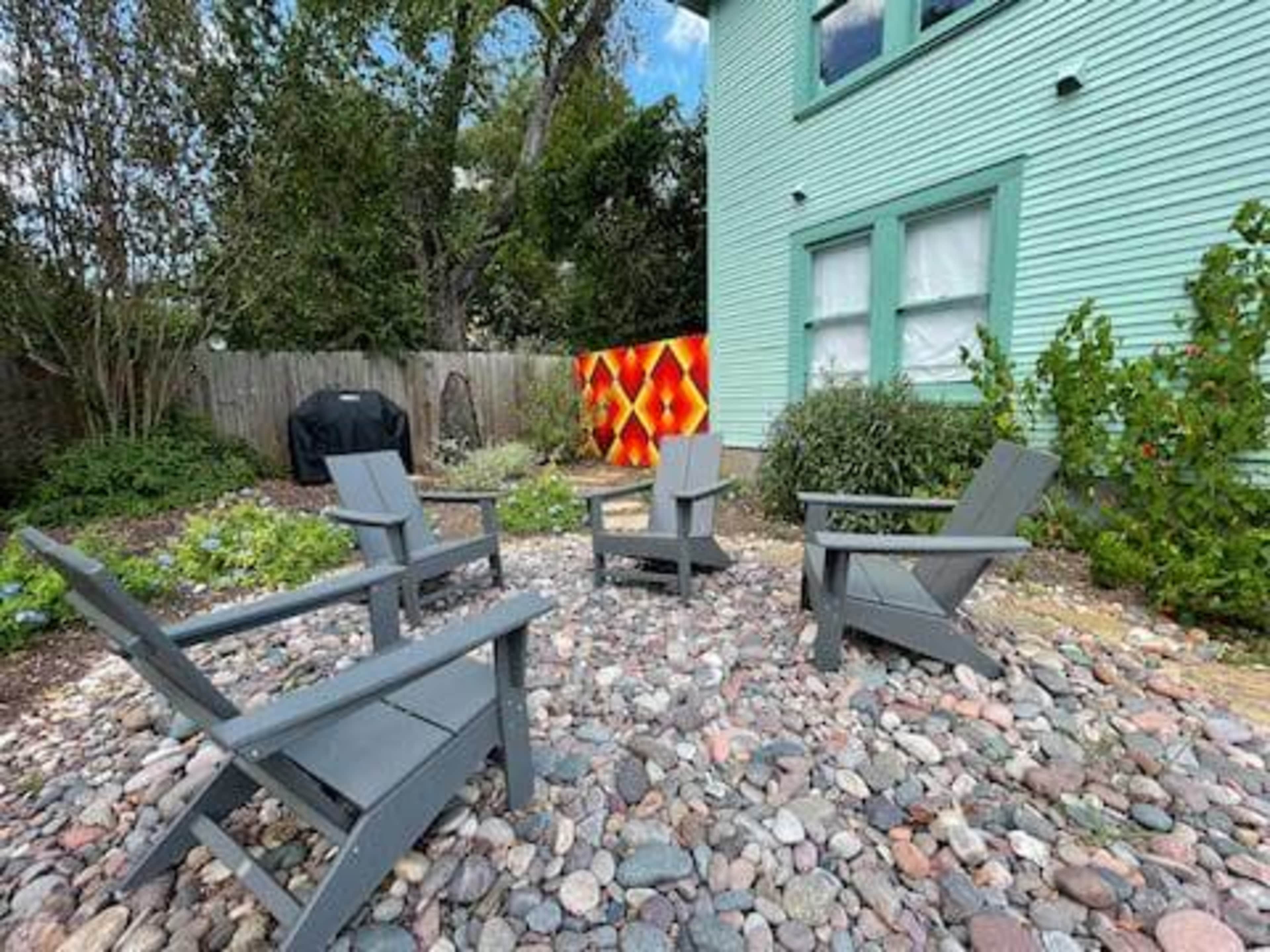 A small outdoor seating area features four gray chairs arranged on a bed of pebbles between a garden and a house with a green exterior.