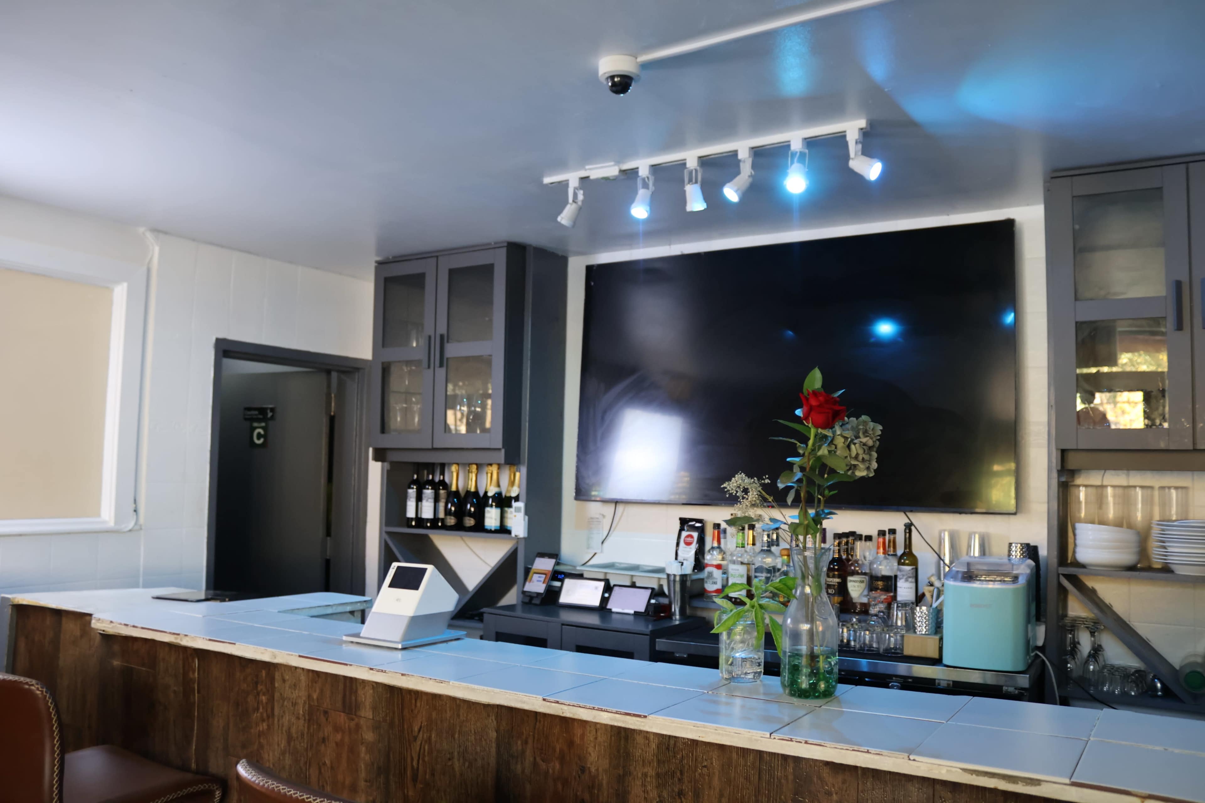 The image shows a bar area with a large television mounted on the wall, shelves stocked with liquor, and a floral arrangement on the countertop.