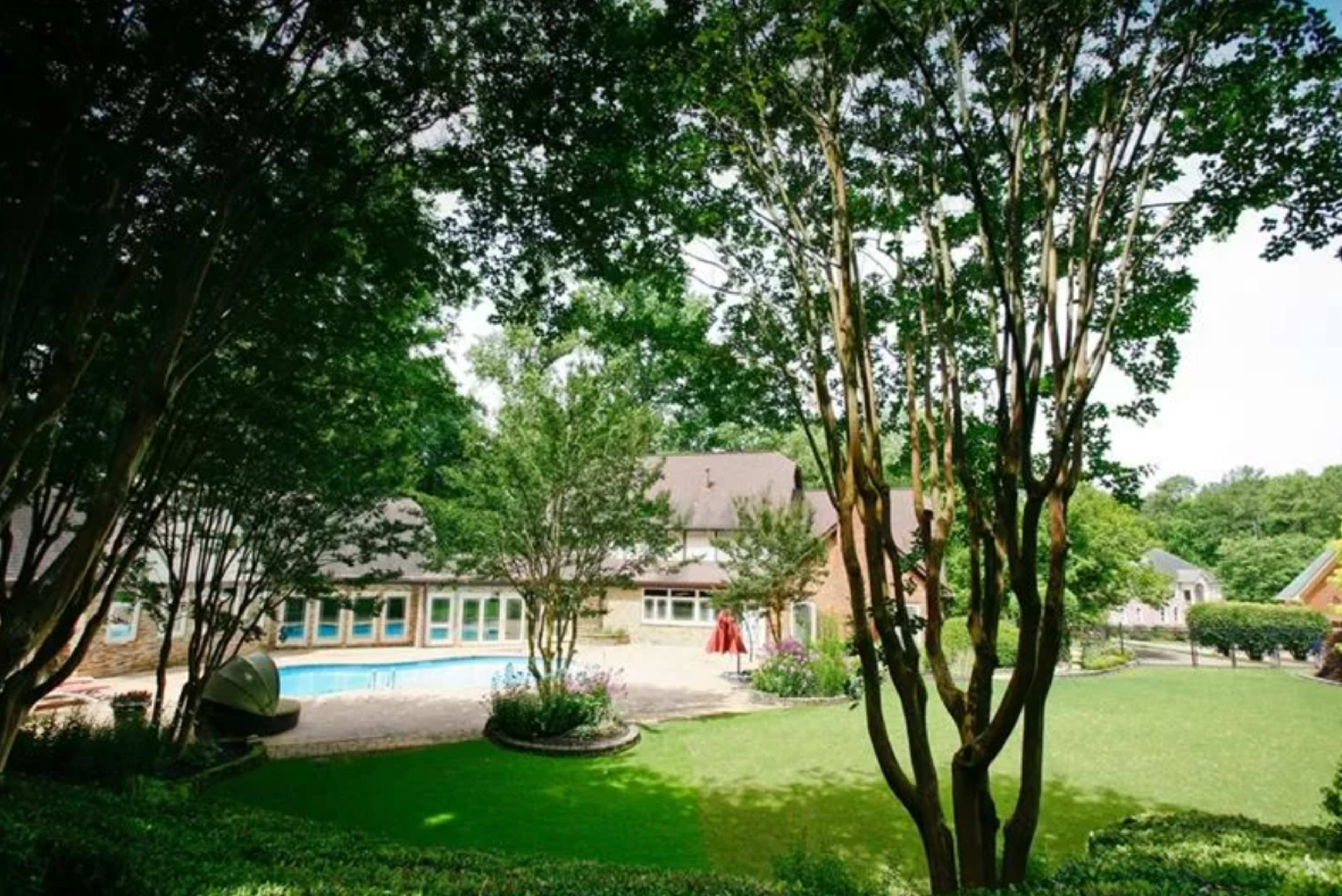 A backyard features a swimming pool surrounded by lush grass and trees, with a house in the background.