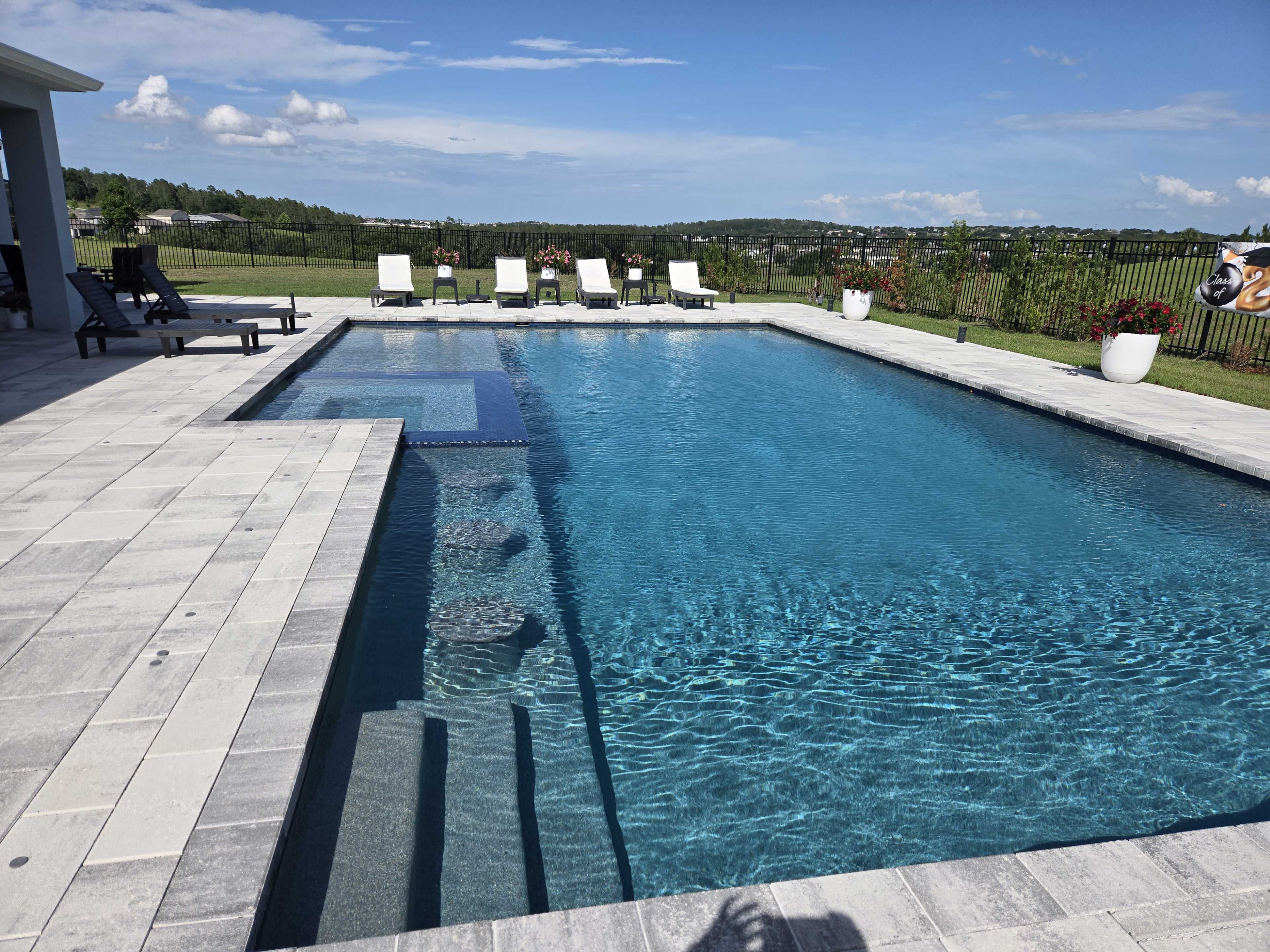 The image shows a large, clear swimming pool surrounded by lounge chairs and greenery under a blue sky.