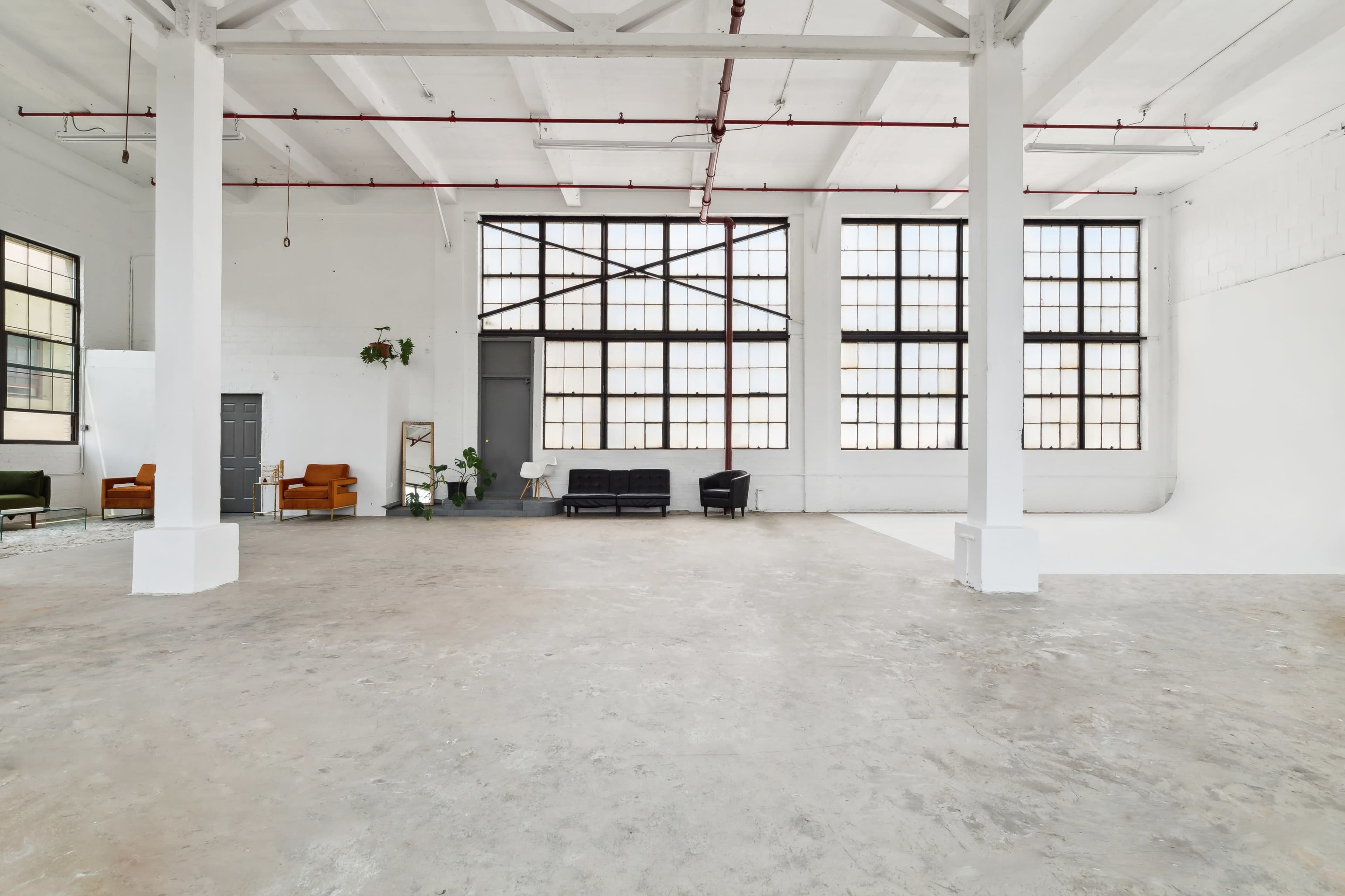 A spacious, empty room with large windows, concrete flooring, and minimal furniture including a black sofa and two armchairs.