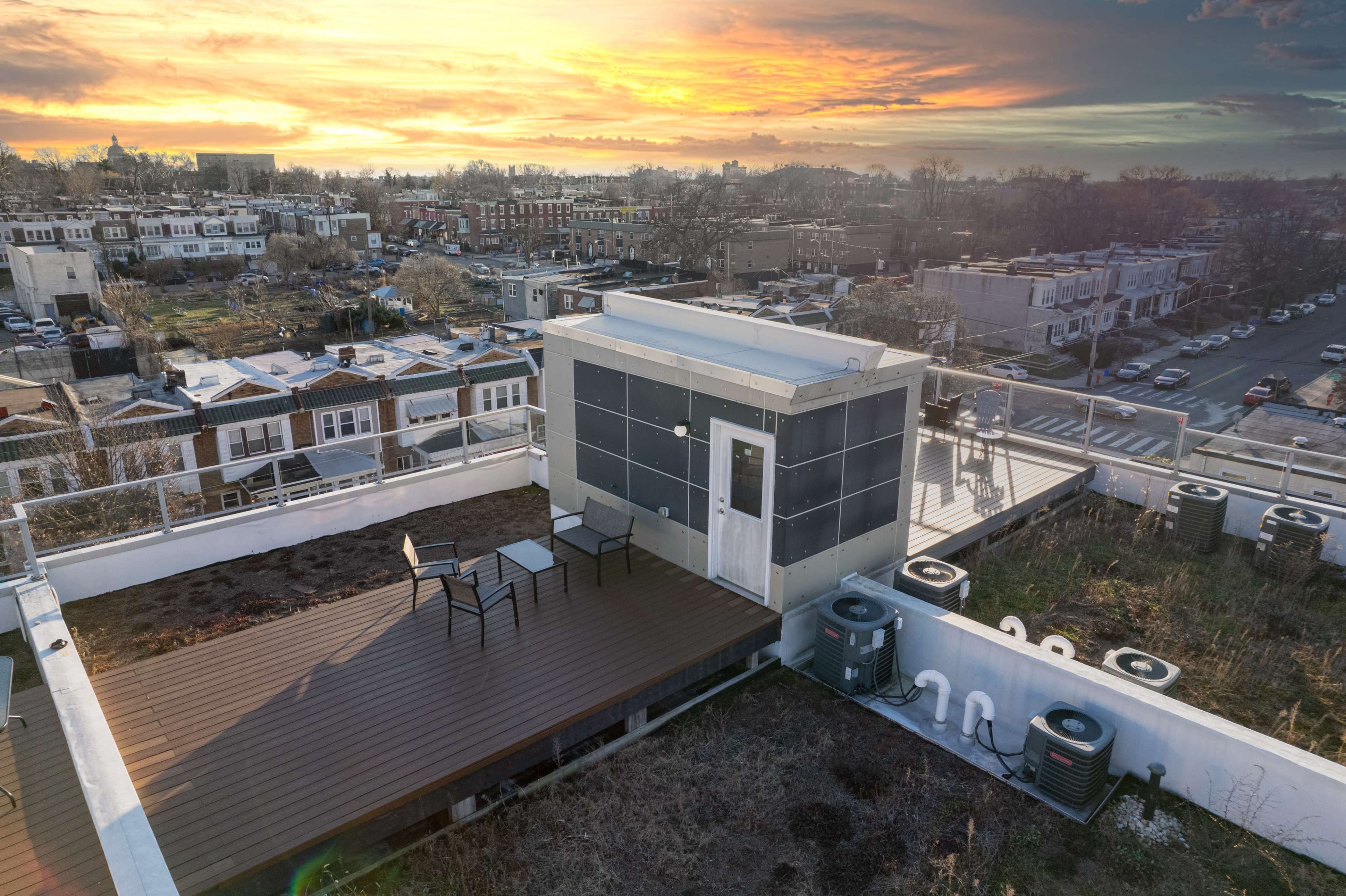 A rooftop deck features two chairs and a table overlooking a neighborhood with houses and a sunset in the background.