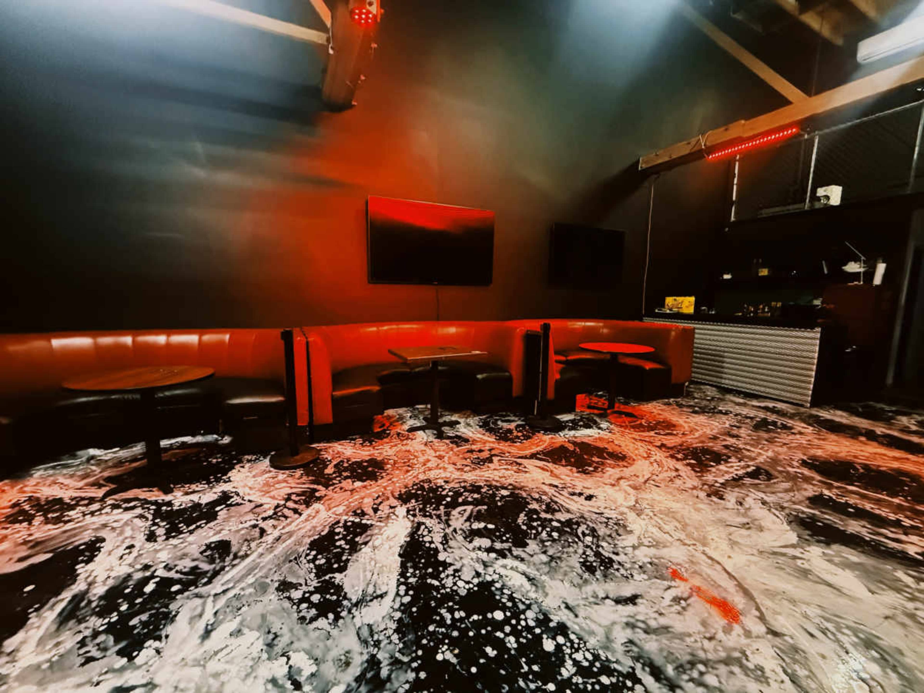 The image shows a dimly lit room with red accents, featuring a wet floor covered in foam and scattered tables and seating.