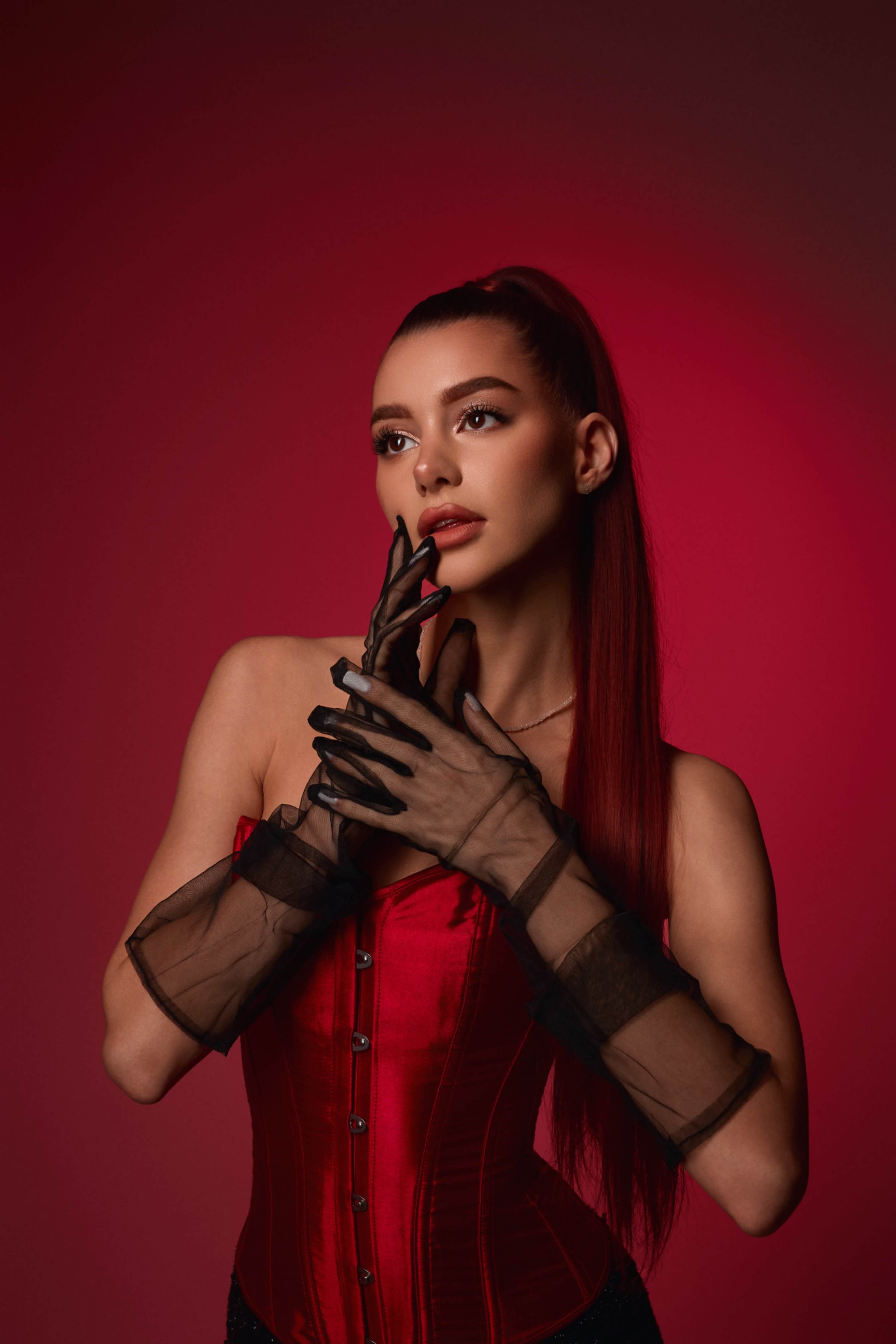 A woman wearing a red corset and black gloves poses against a deep red background.