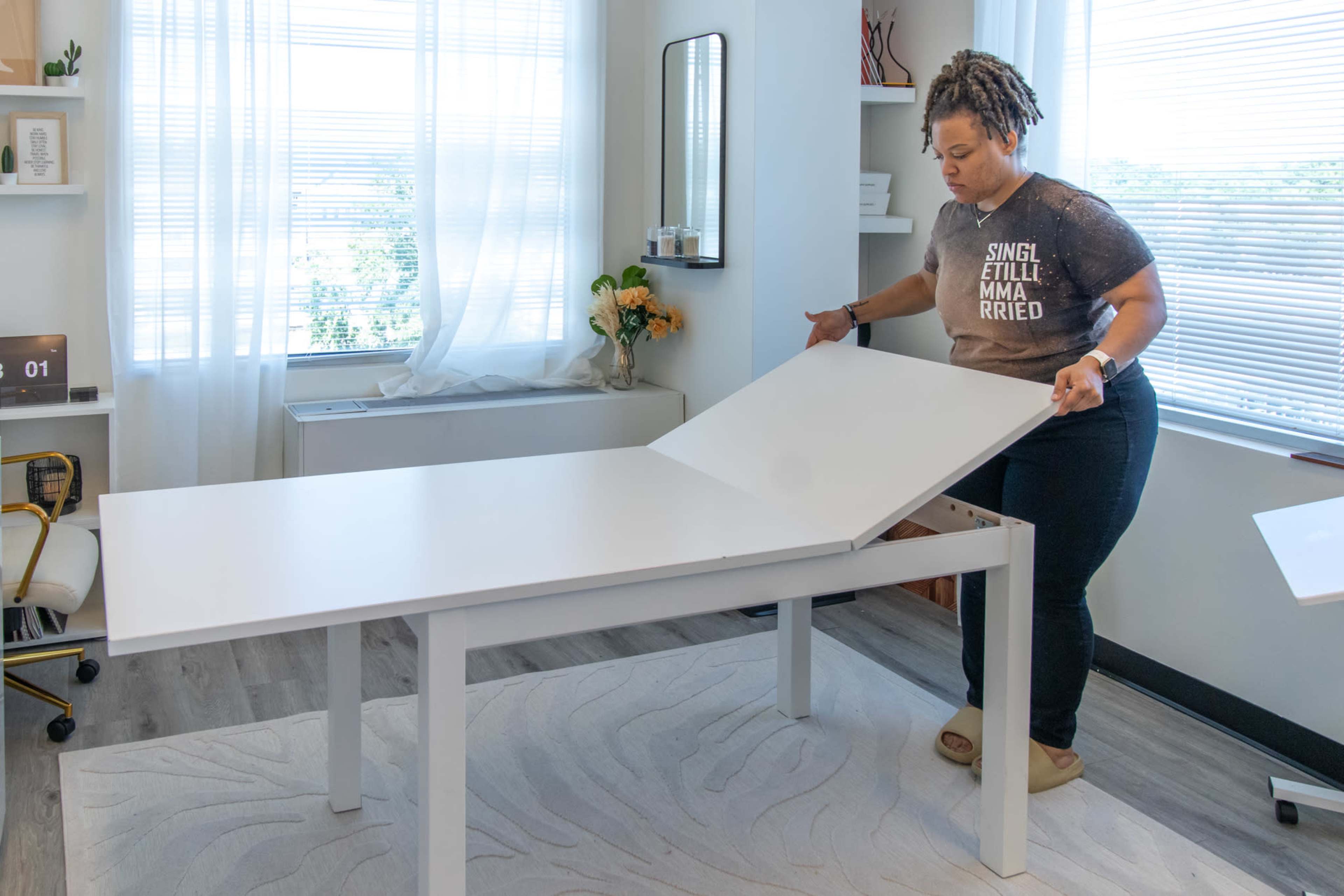 A woman adjusts a white, extendable table in a brightly lit room with large windows.