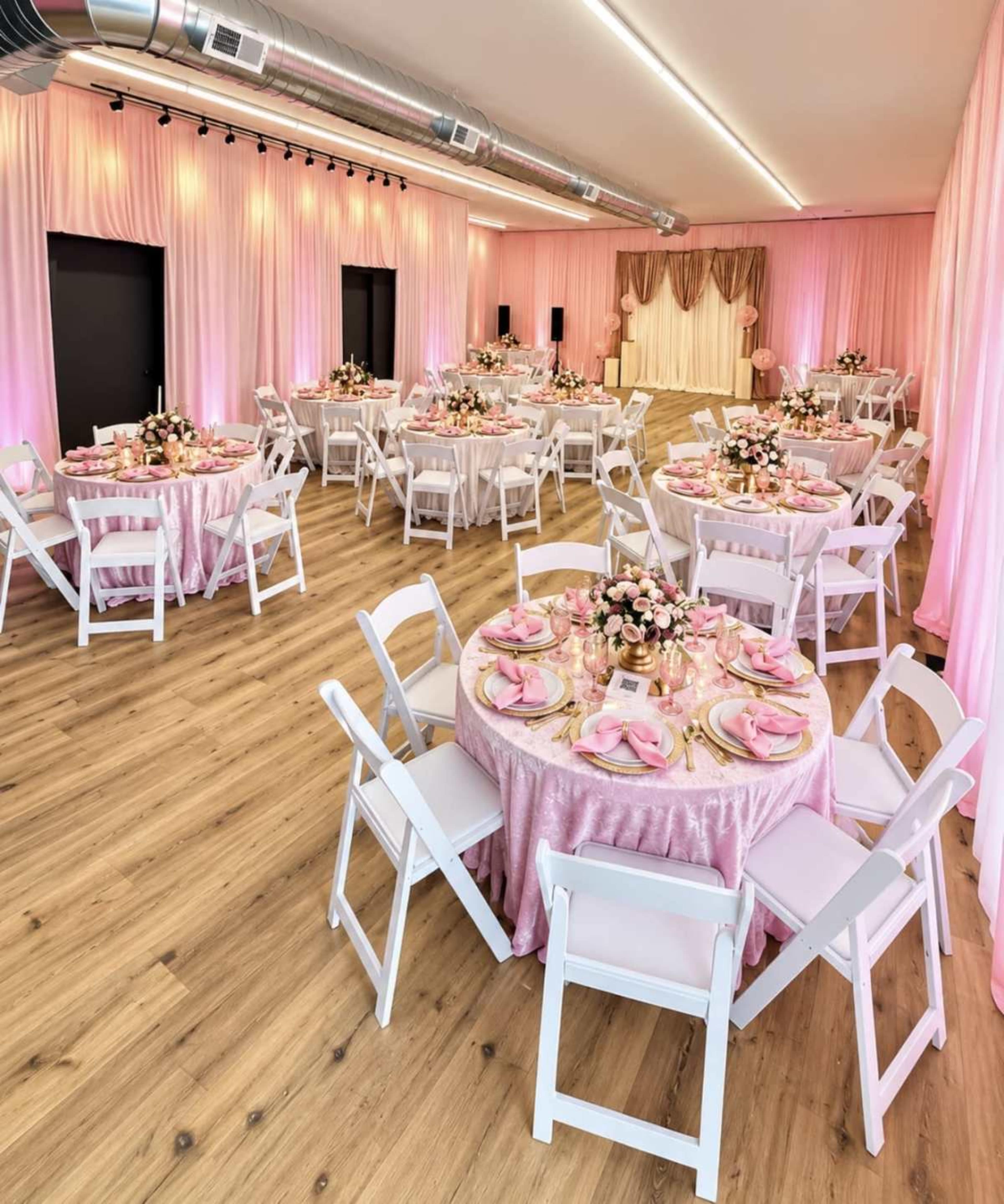 The image shows an elegantly arranged event space with round tables covered in pink tablecloths, adorned with floral centerpieces and white chairs, all set against soft pink drapery on the walls.