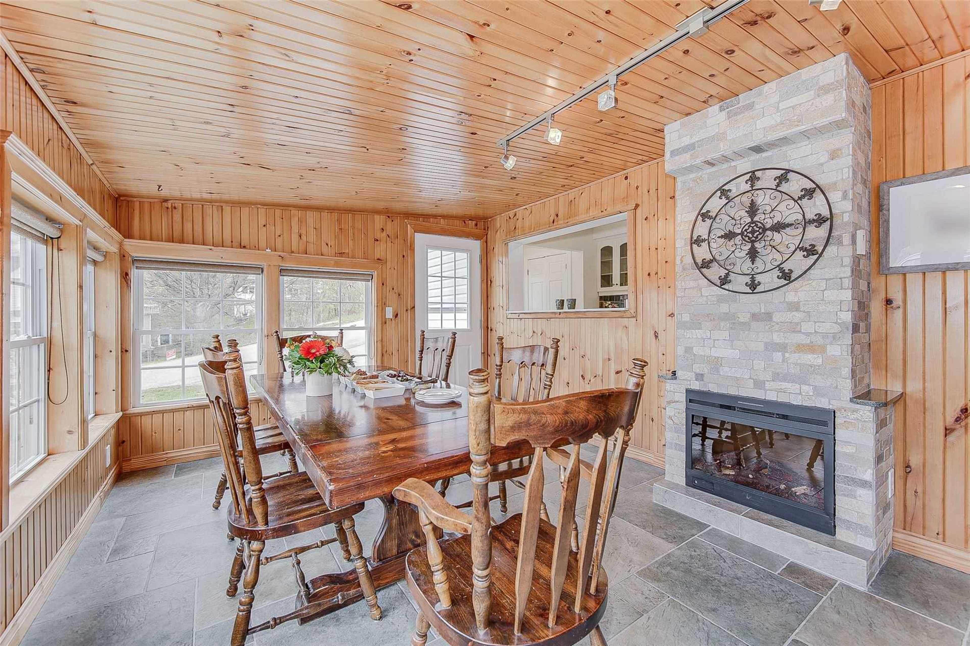 The image shows a wooden dining area with a long table, surrounded by chairs, and a stone fireplace on one wall.