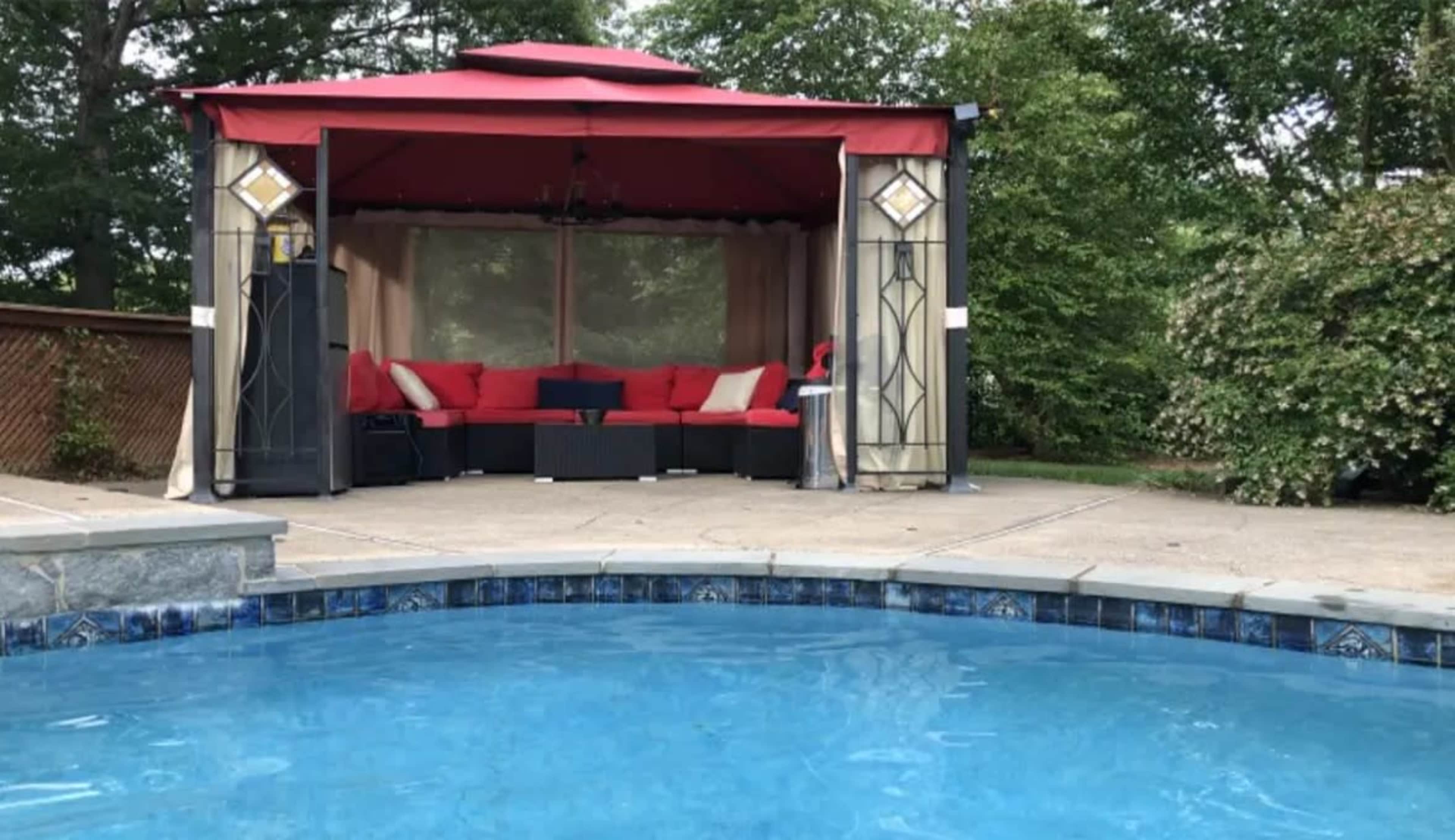 A backyard scene featuring a swimming pool in the foreground and a gazebo with red cushions and a canopy in the background.
