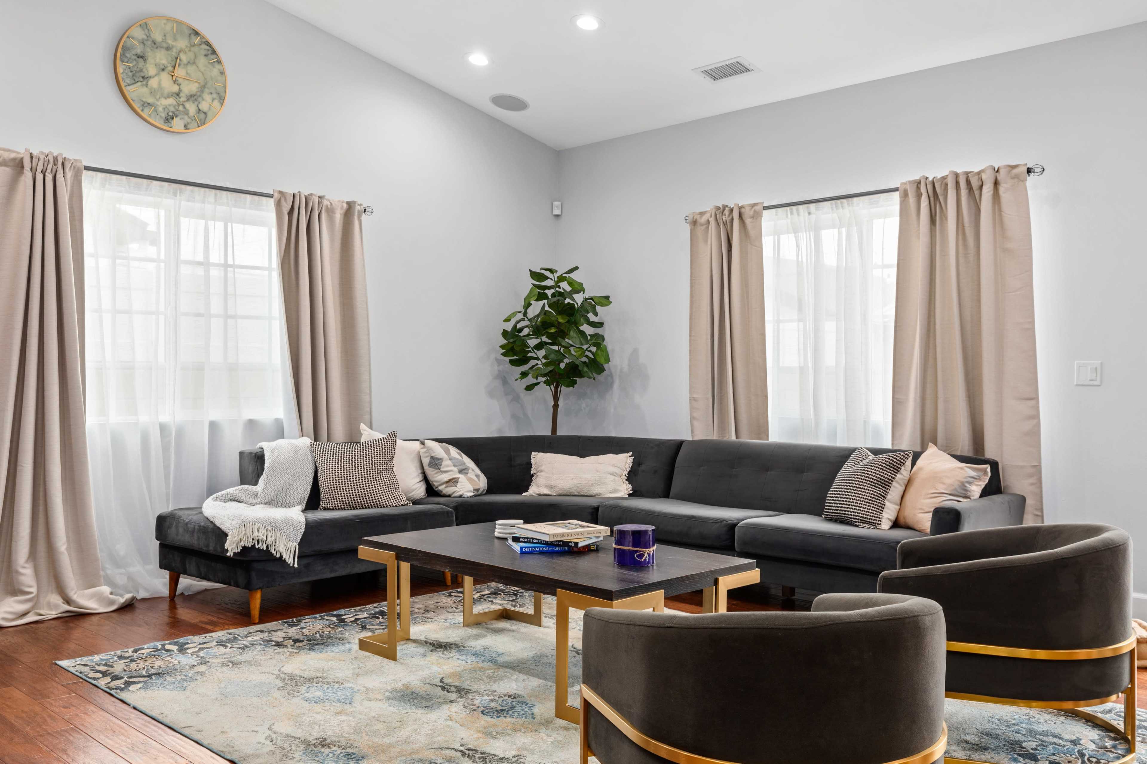 A modern living room features a grey sectional sofa, two circular chairs, a coffee table, and large windows with light curtains.