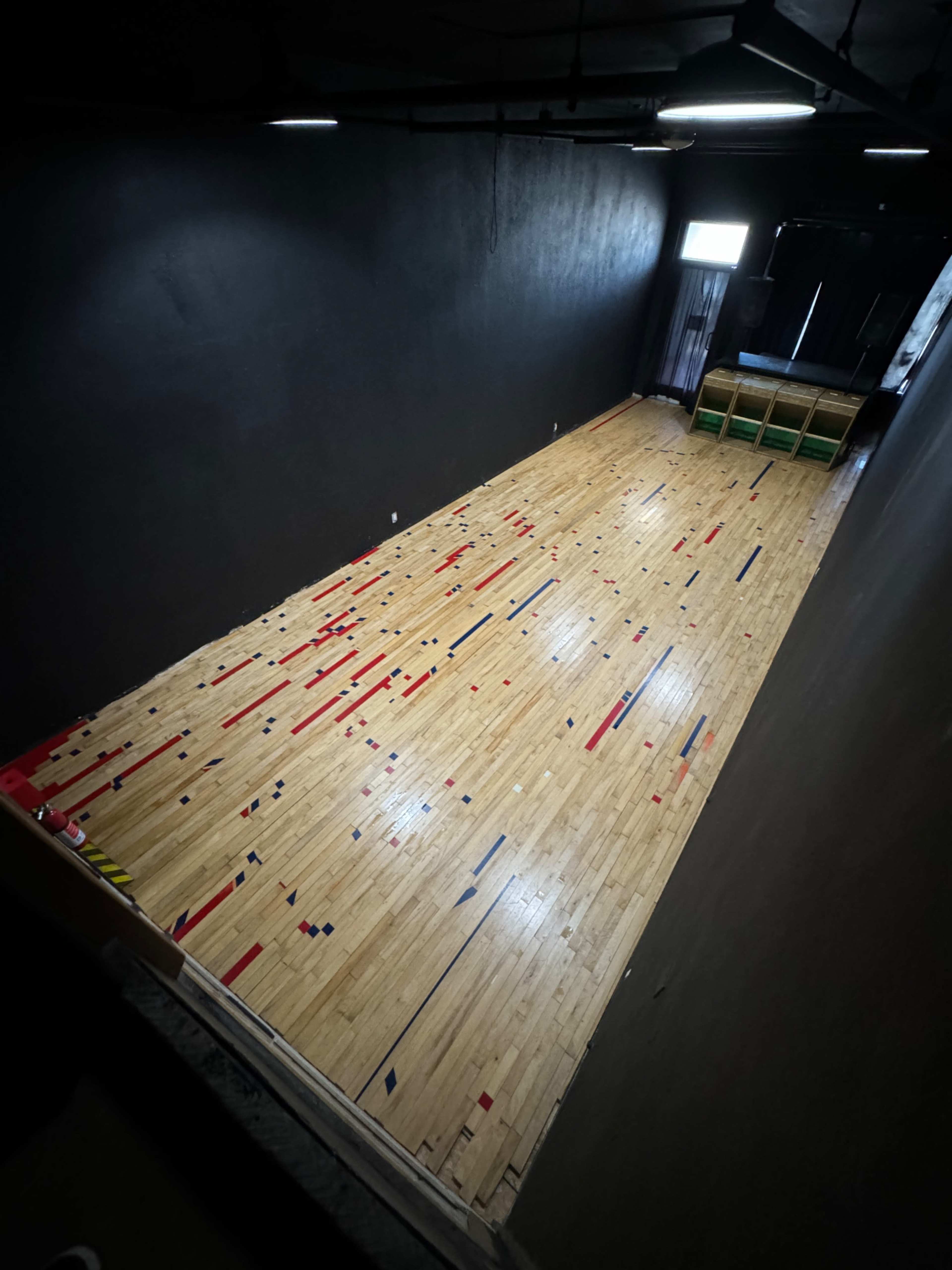 The image shows a long, narrow room with a polished wooden floor featuring a pattern of red, blue, and yellow lines, illuminated by overhead lights.