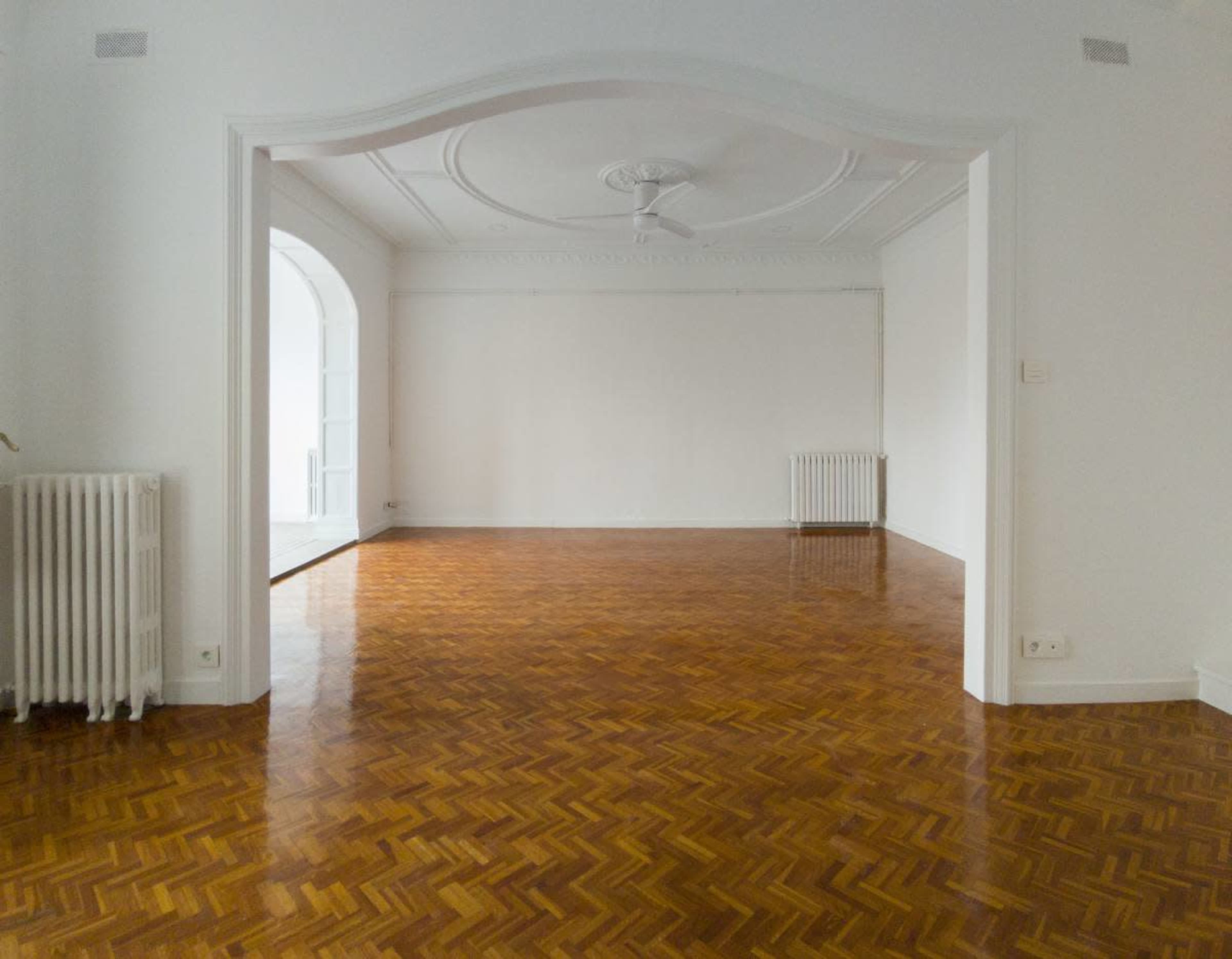The image shows a spacious, empty room with polished wood flooring, a ceiling fan, and large doorways leading to other areas.