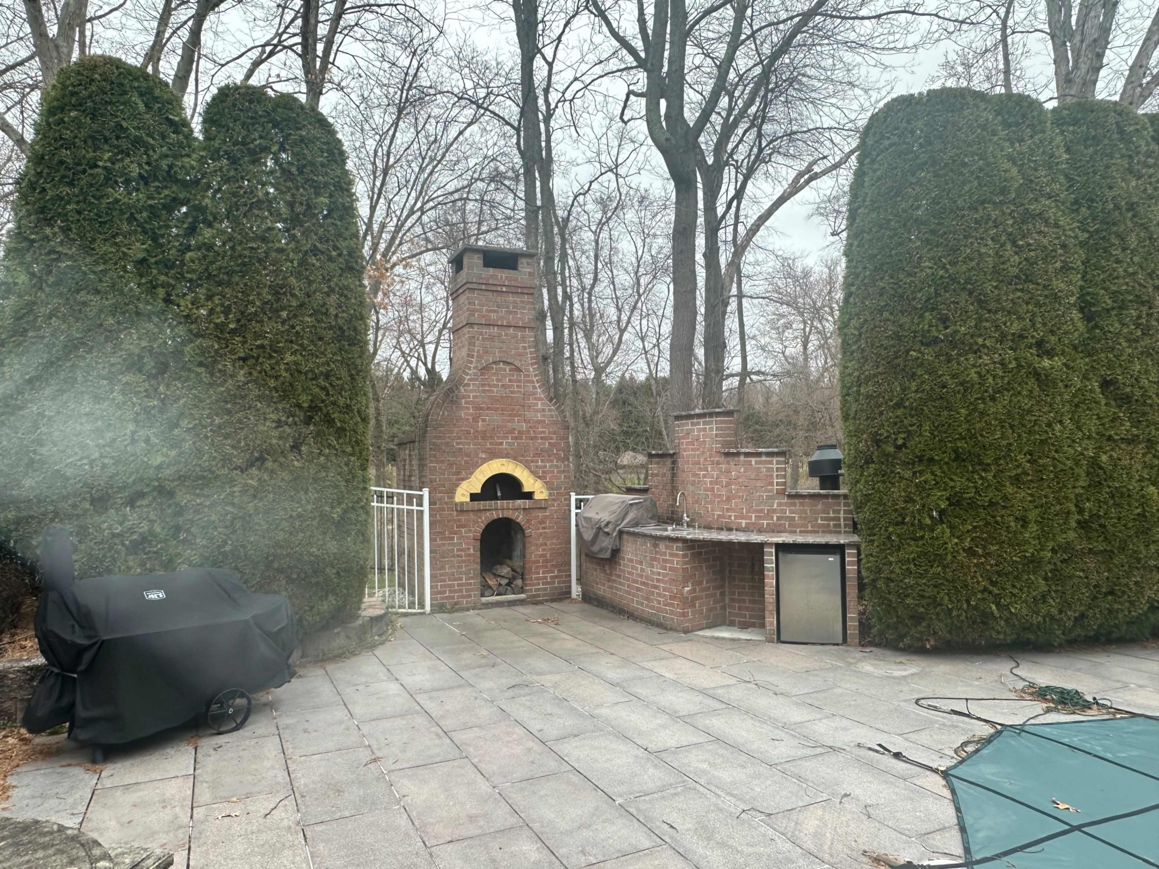 A brick outdoor fireplace surrounded by tall evergreen shrubs, with a grill and a stone patio visible in the scene.