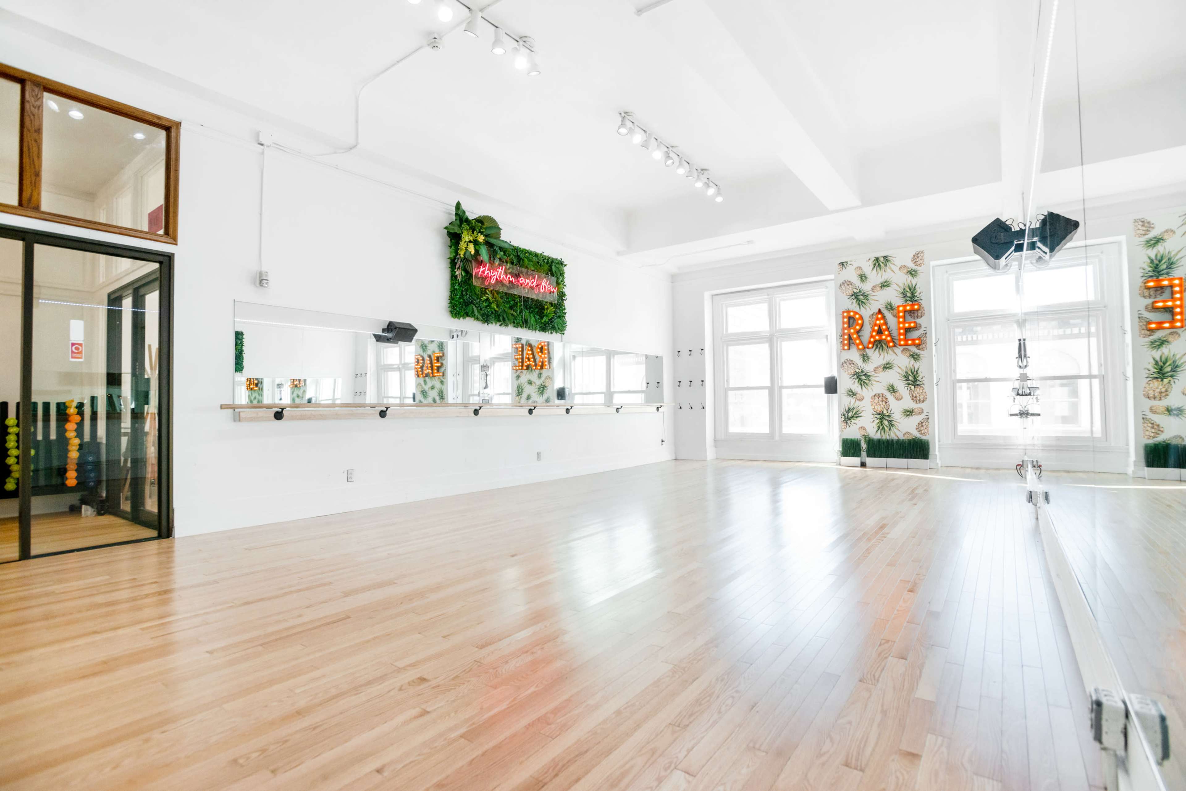 A bright, spacious dance studio with wooden flooring, large mirrors, and decorative wall elements featuring plants and illuminated letters.