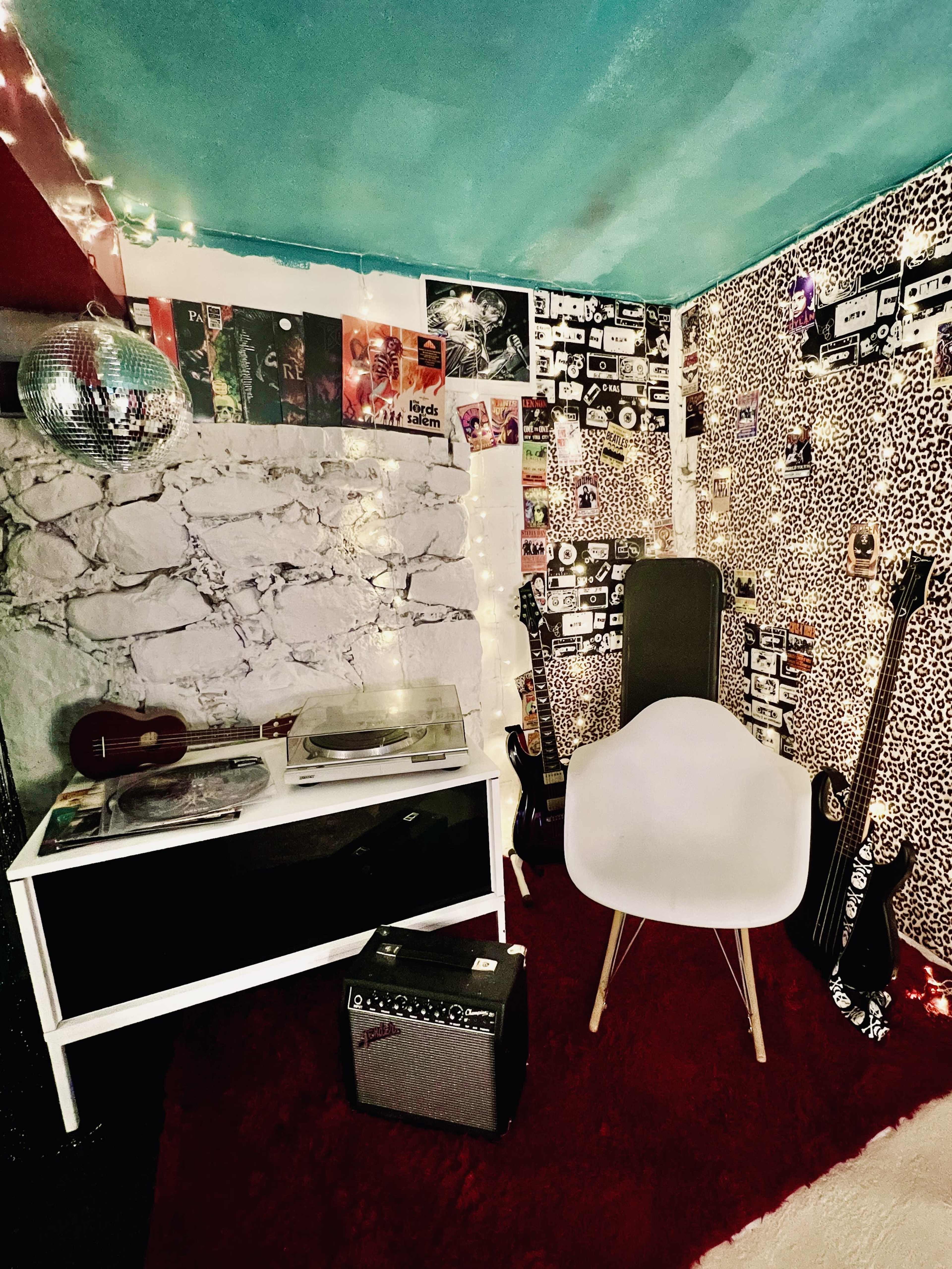 The image shows a cozy corner of a music-themed room featuring a white chair, a record player, an amplifier, and guitars against a backdrop of patterned wallpaper and string lights.