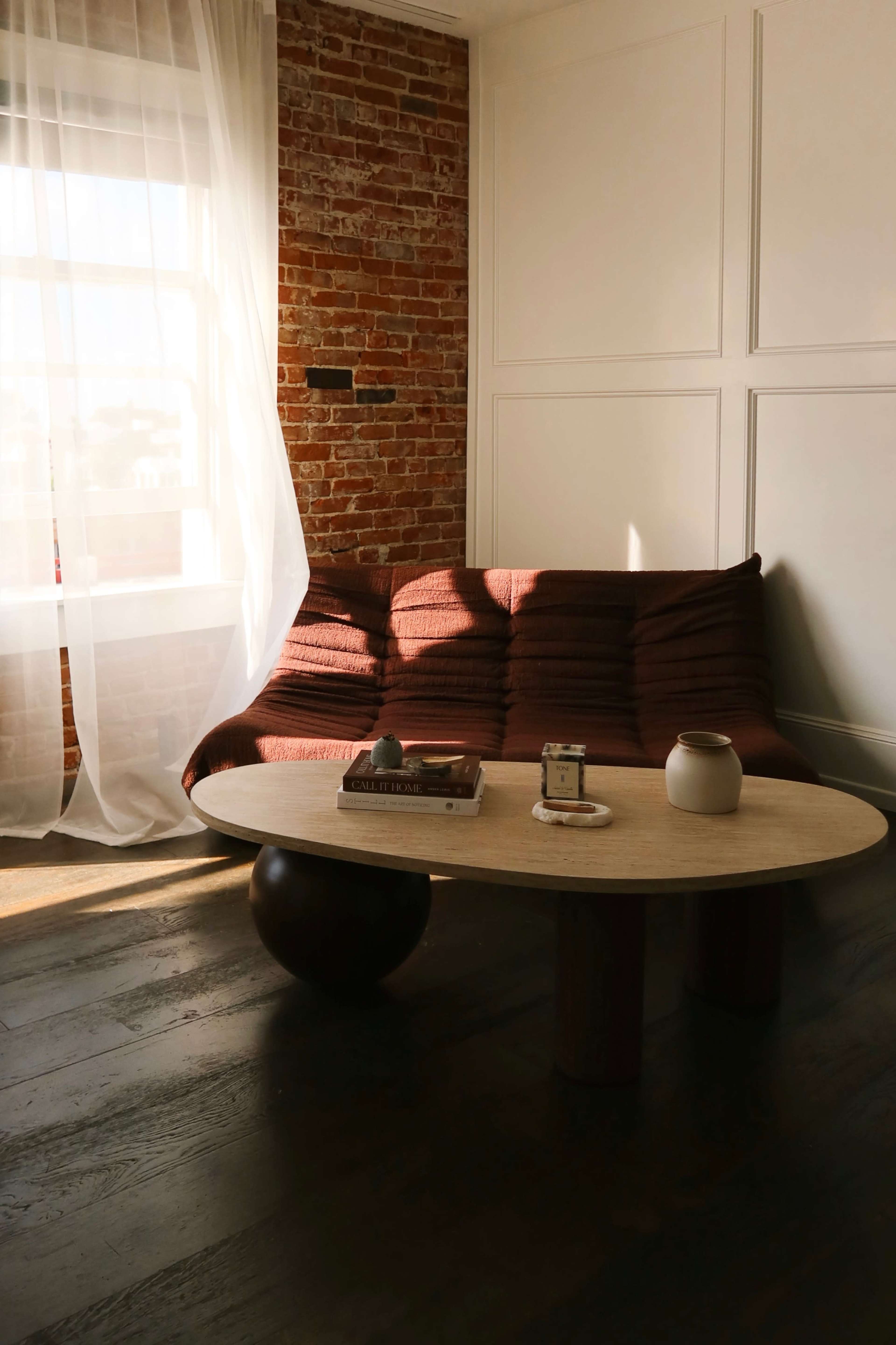 The image features a minimalist living space with a brown sofa and a round wooden coffee table, illuminated by natural light from a large window.