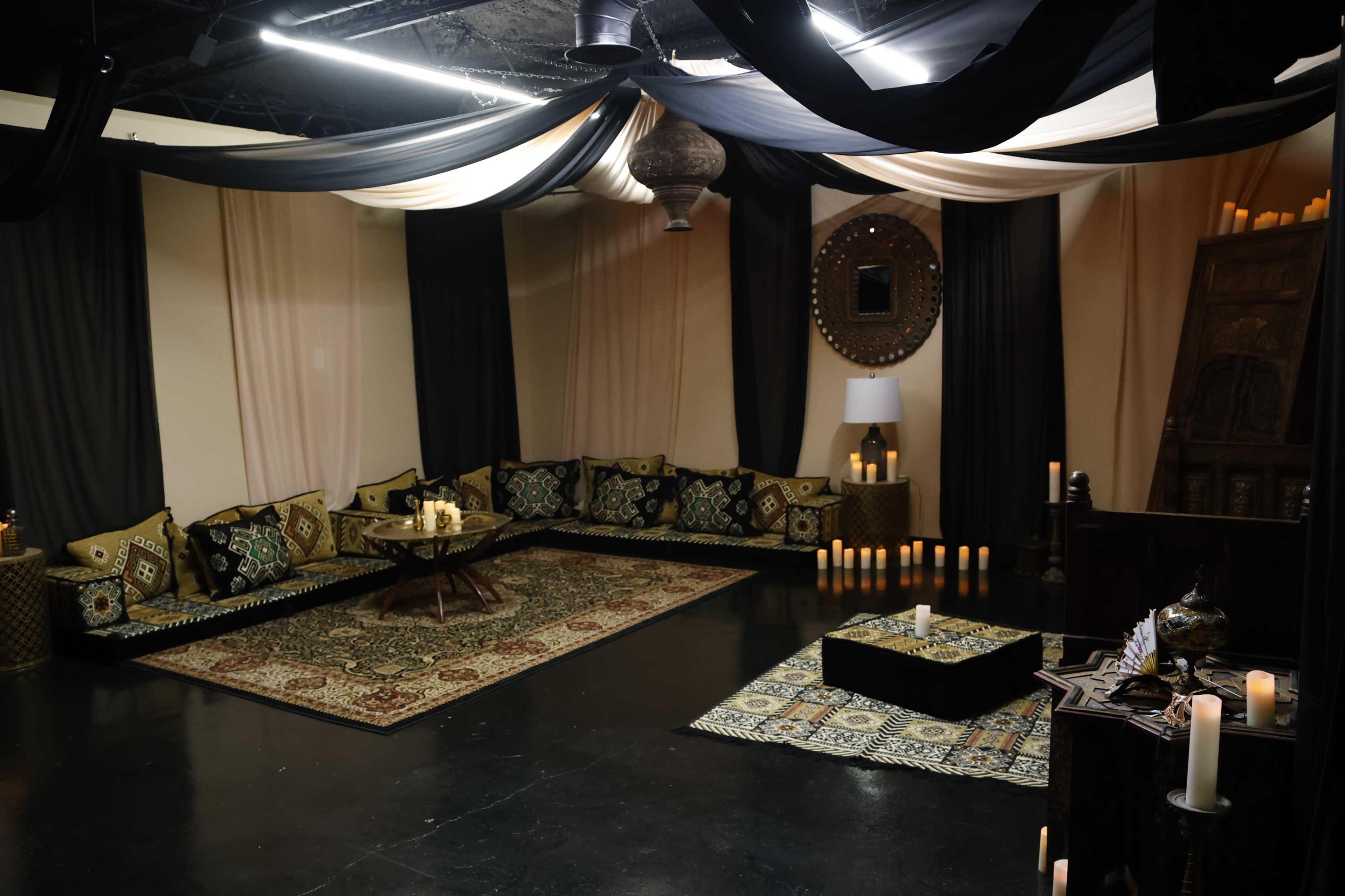 A dimly lit room decorated with dark drapes, patterned cushions along a low table, and candles of varying heights arranged around a central area.