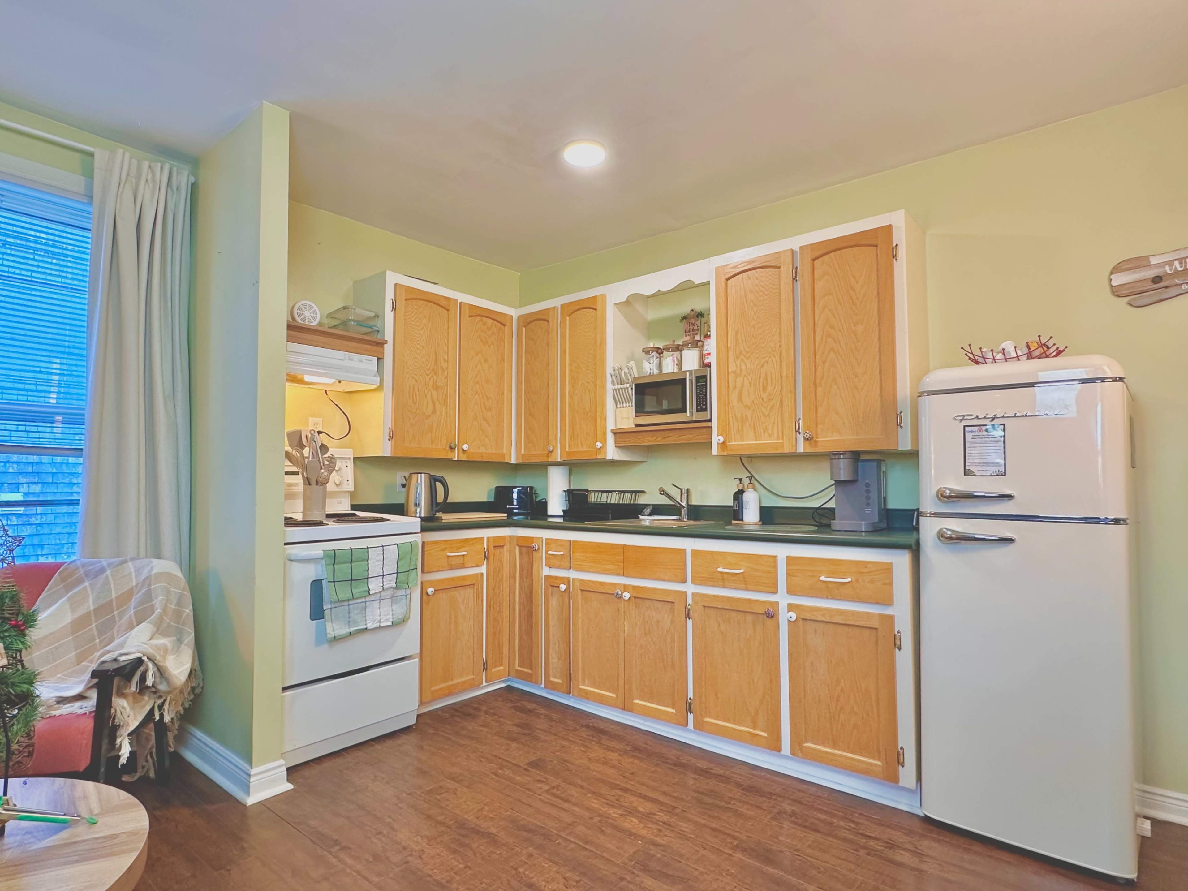 The image shows a kitchen with wooden cabinets, a refrigerator, a stove, and various appliances set against a light green wall.