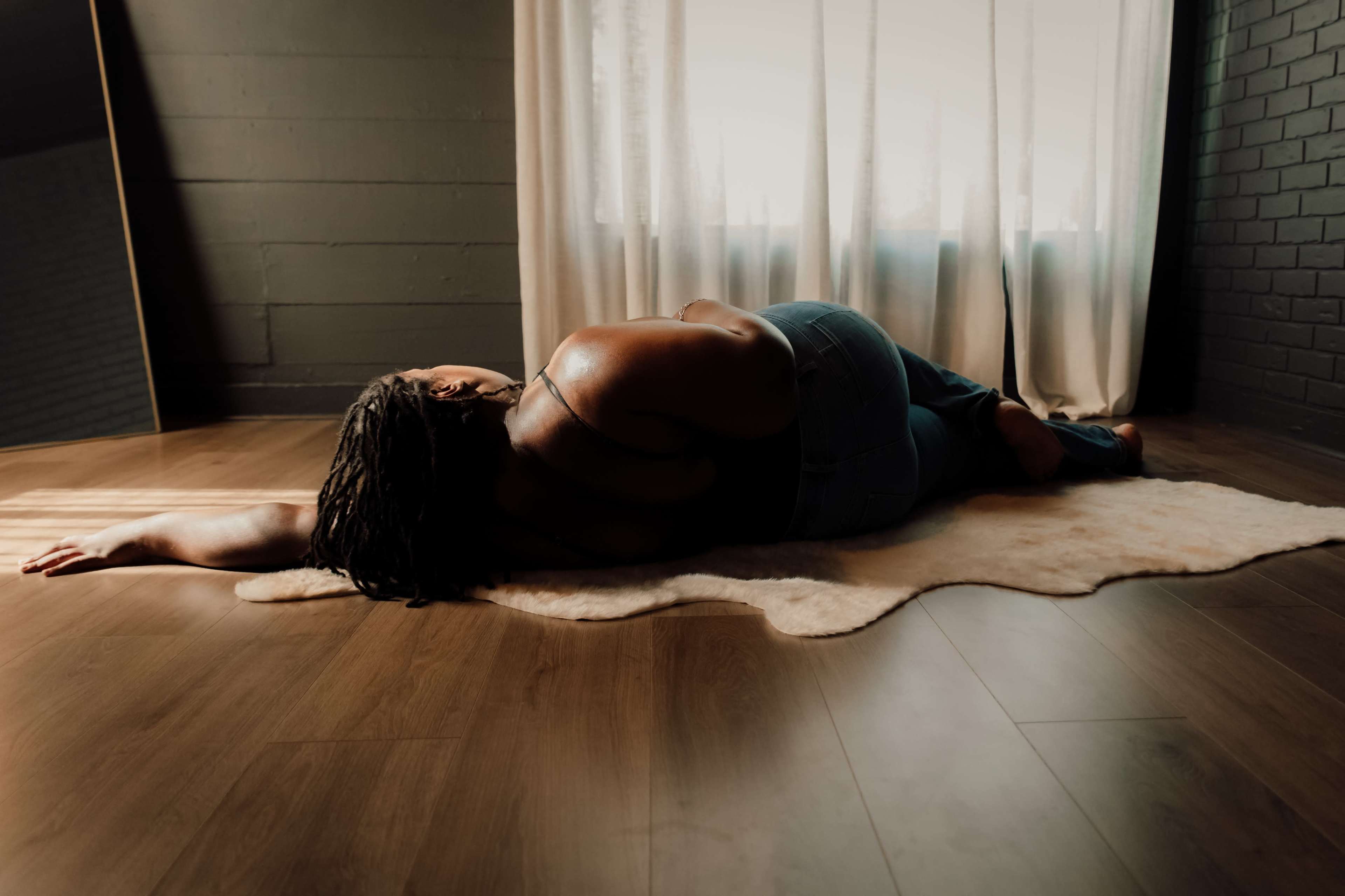 A person with long hair lies on a rug, facing away from a window with light streaming through sheer curtains.