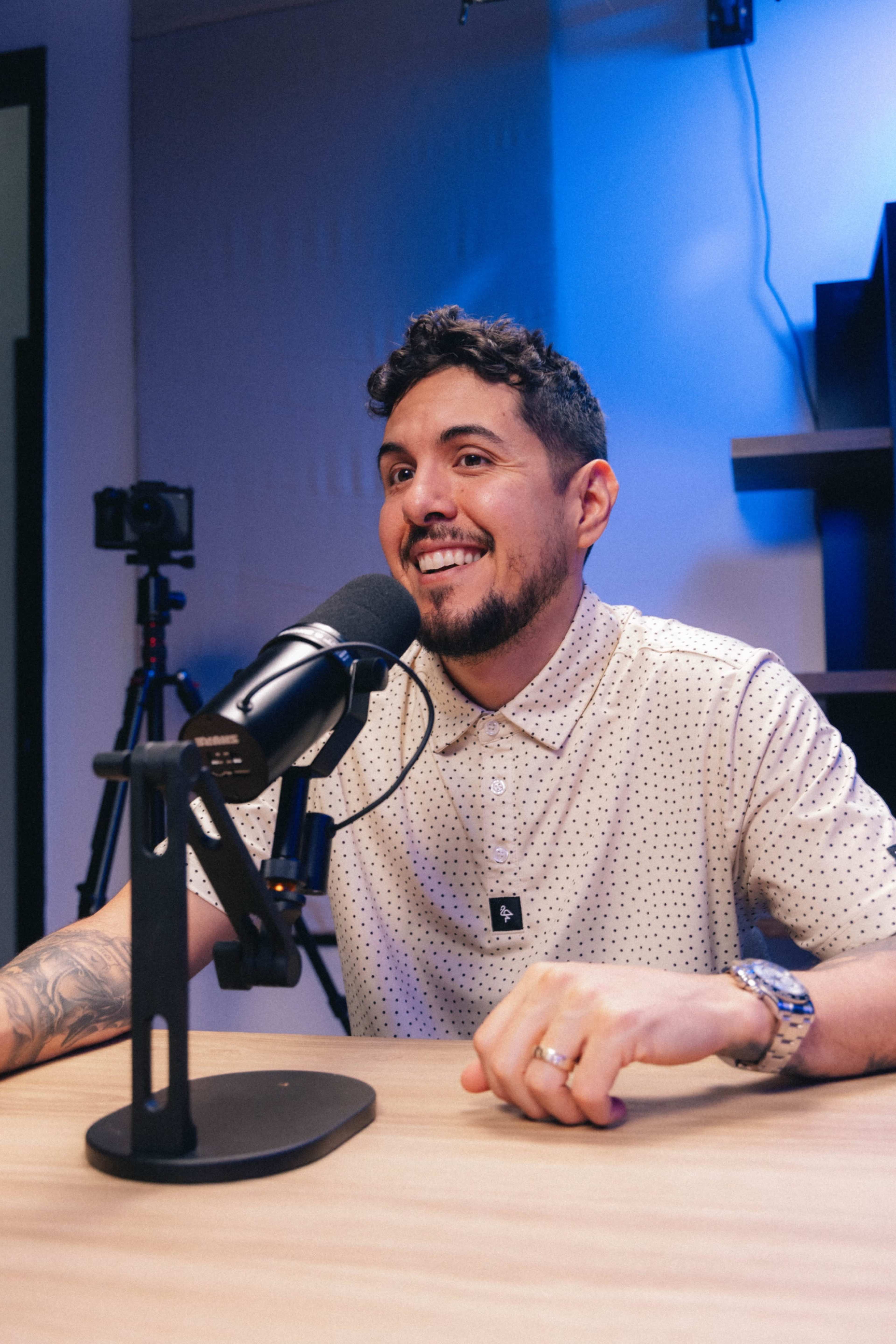 A man with a neatly styled beard sits at a table with a microphone in front of him, smiling during a podcast recording.