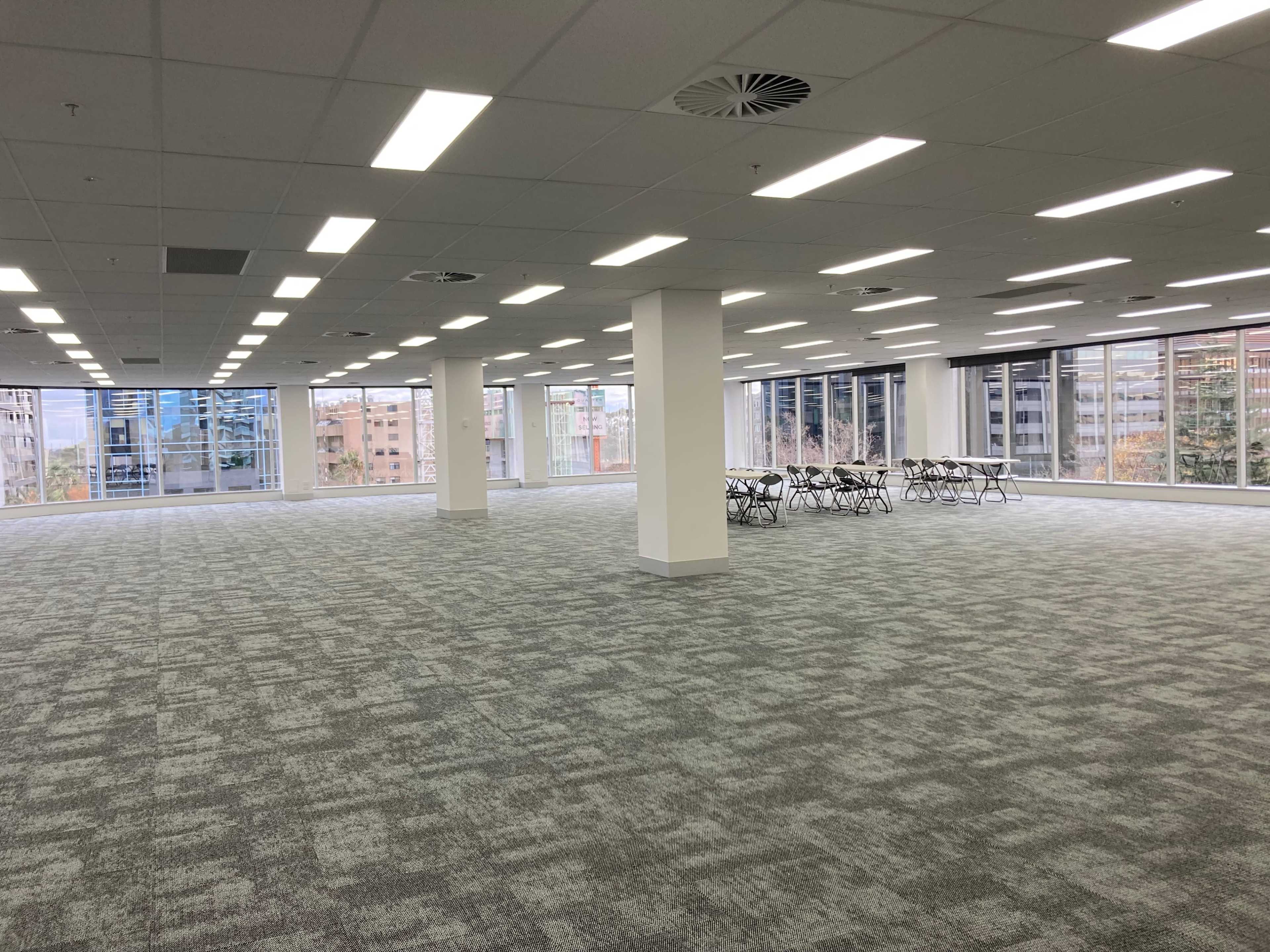 An empty office space features large windows, modular carpet tiles, and rows of foldable tables arranged neatly in the corner.