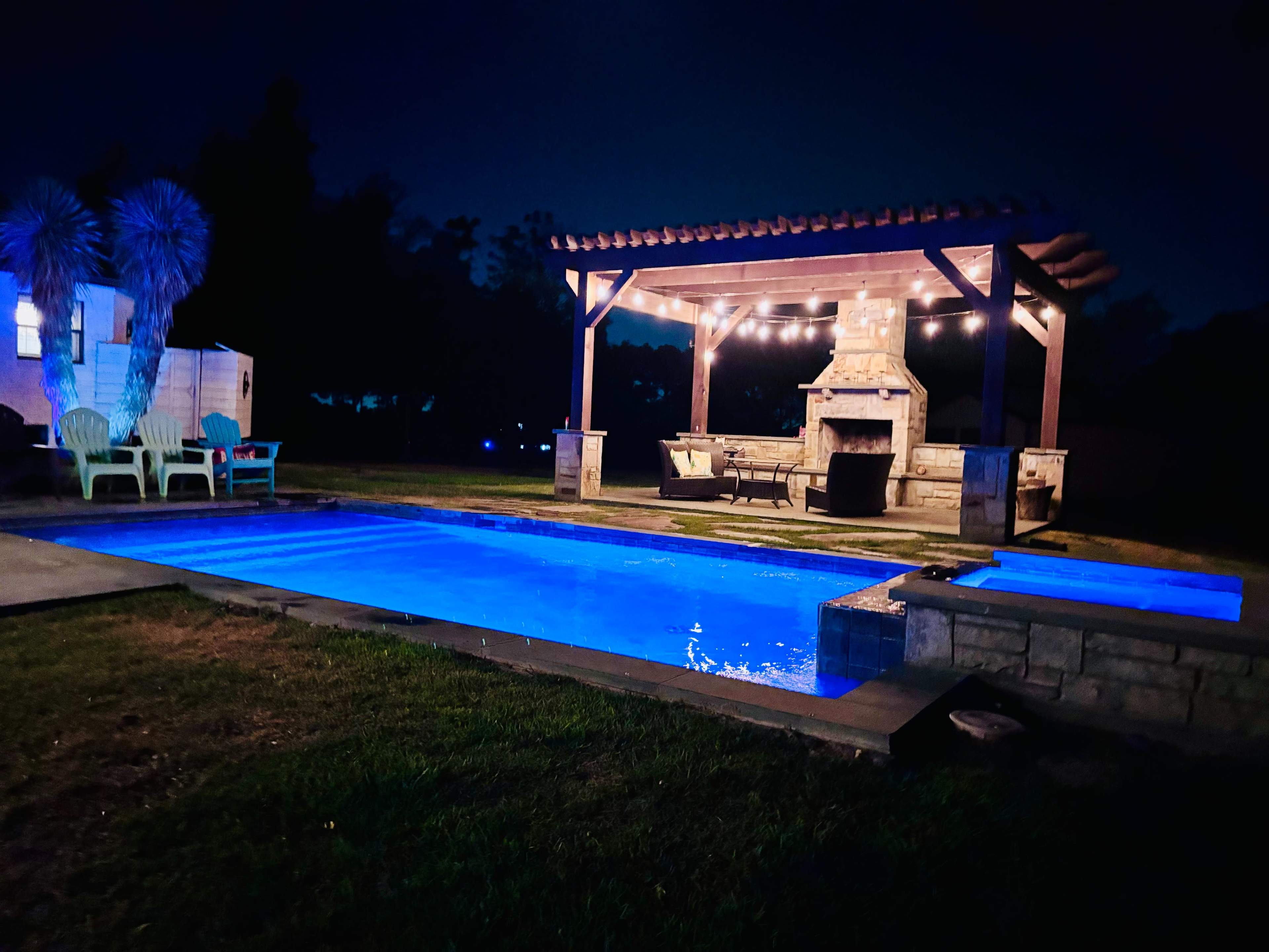 A well-lit outdoor space features a blue-tinted swimming pool and a wooden pergola with seating around a stone fireplace.