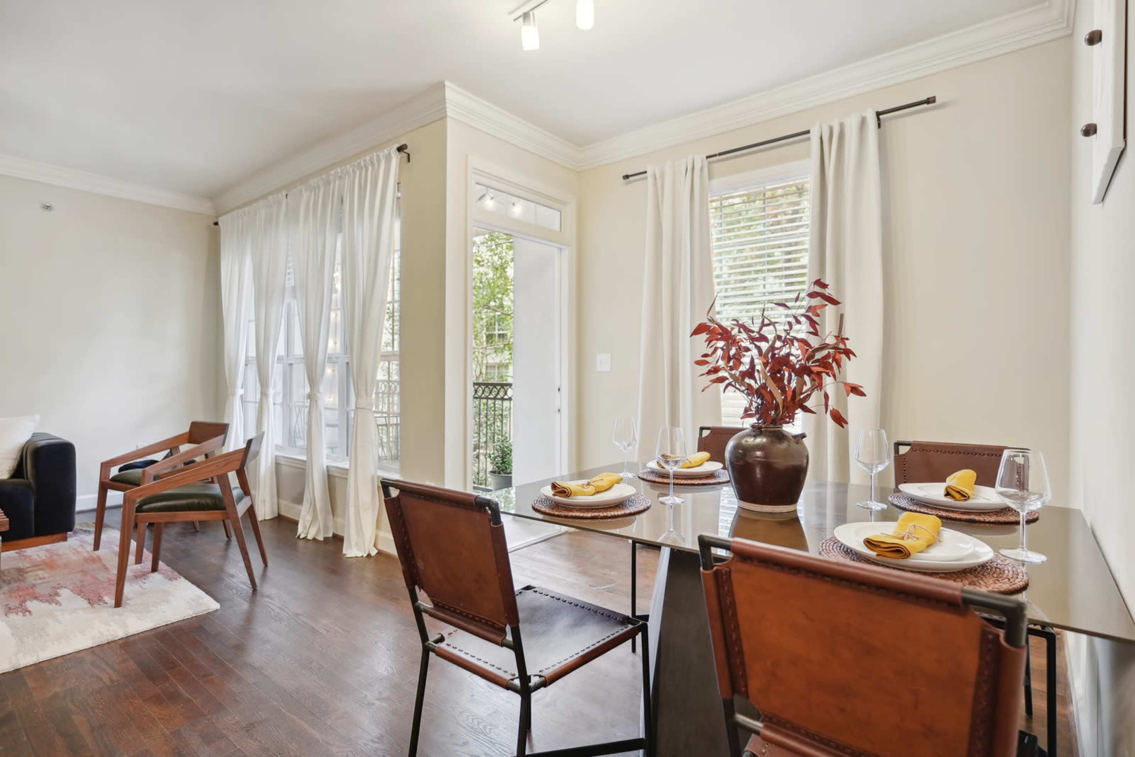 A dining area features a glass table set for four, with a decorative vase and flowing curtains by the windows.