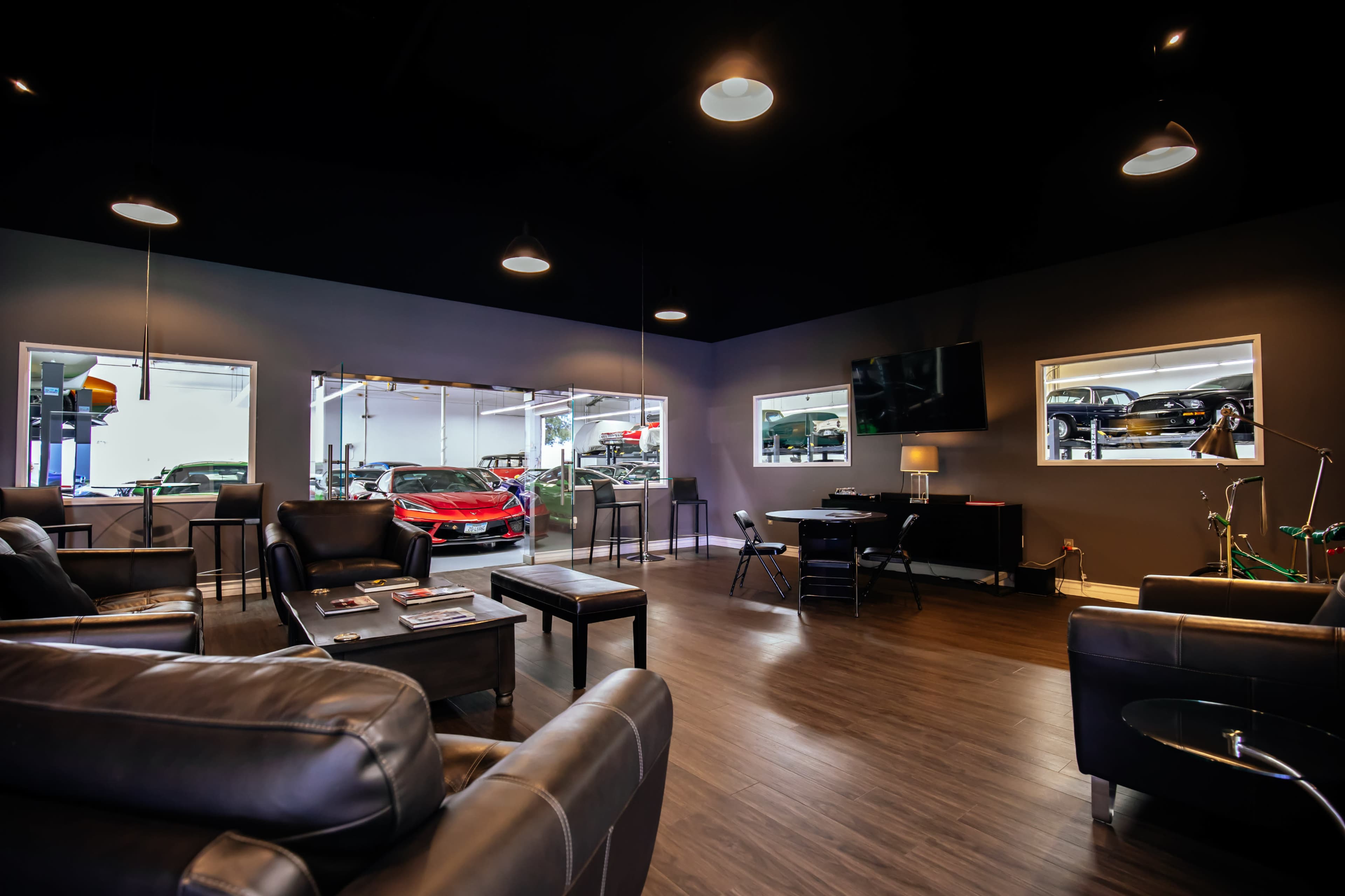 The image shows a modern waiting area with comfortable seating, a television, and large windows overlooking a car service area.
