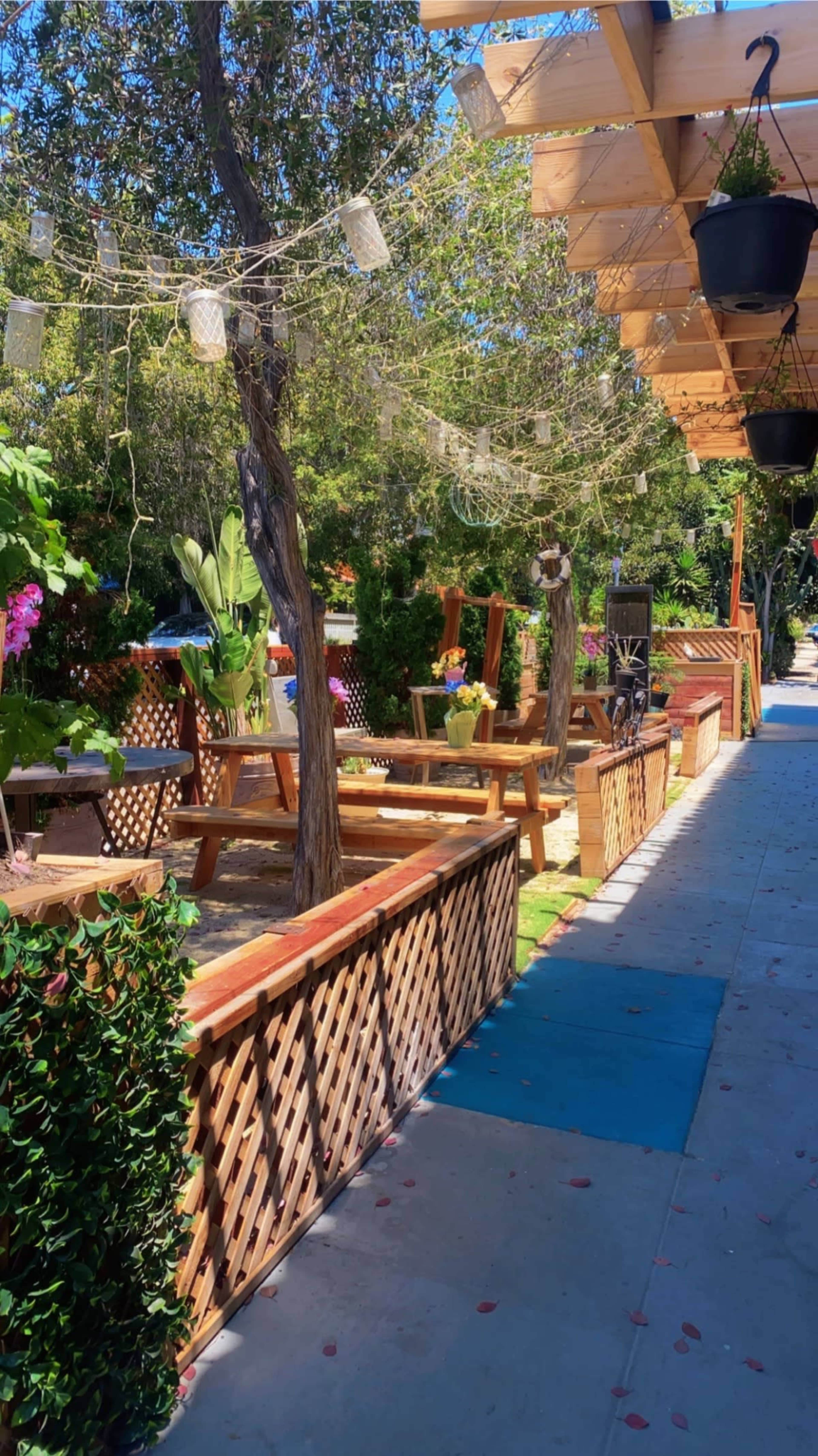 A sunny outdoor seating area with wooden benches and tables, surrounded by greenery and string lights overhead.