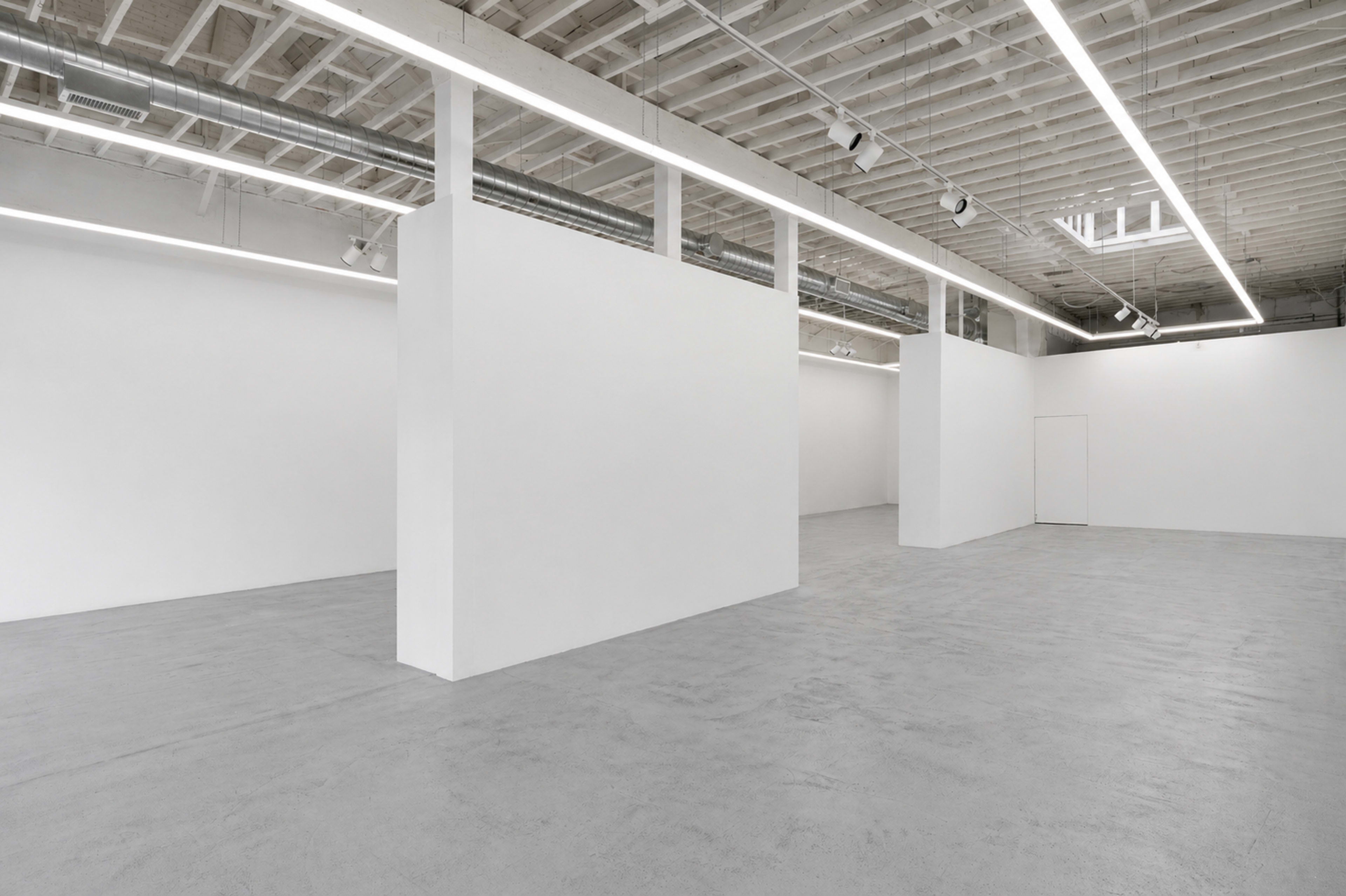 The image shows a stark, empty gallery space featuring white walls, polished concrete flooring, and overhead lighting.