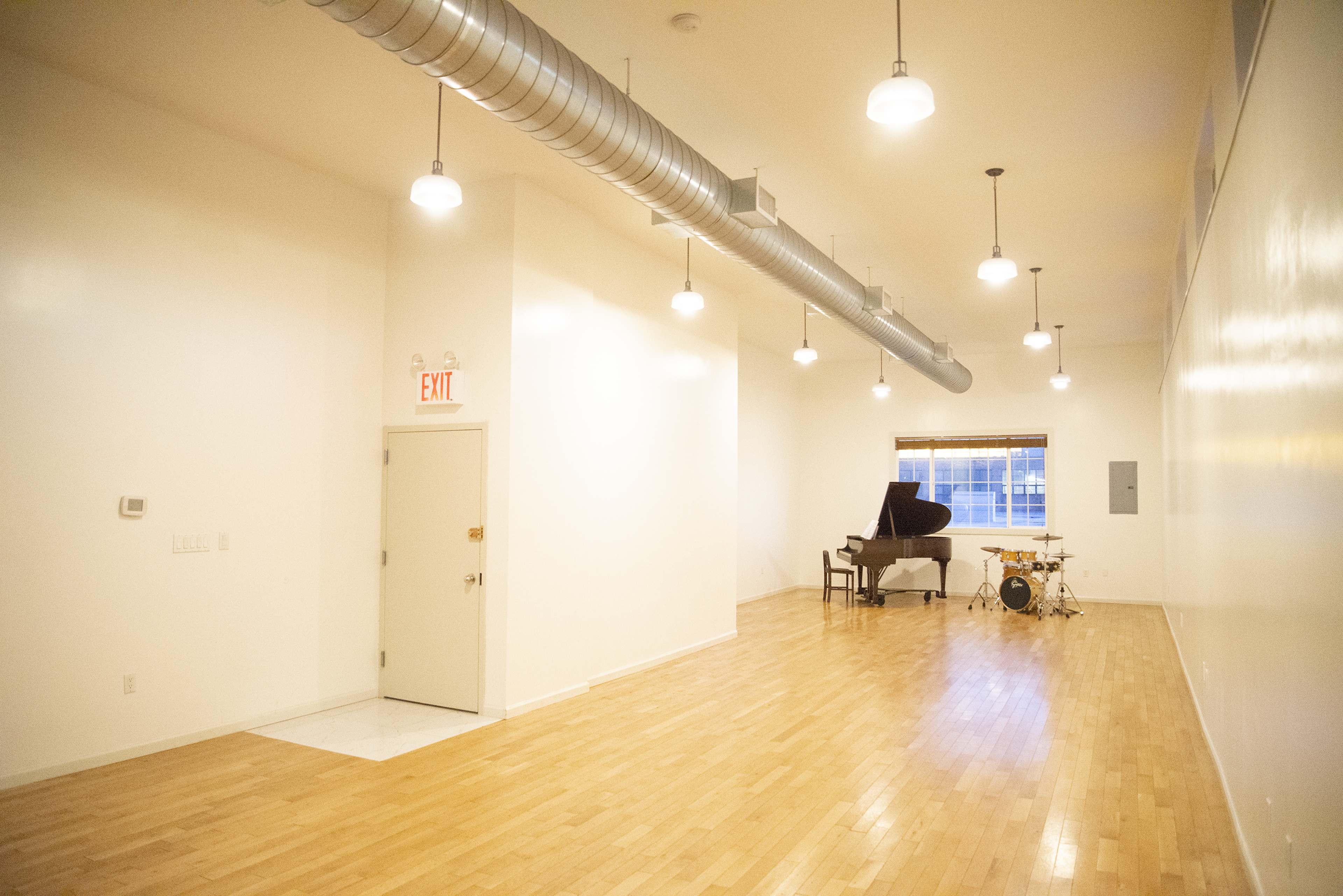 A spacious, well-lit music studio featuring a grand piano and a drum set against a white wall.