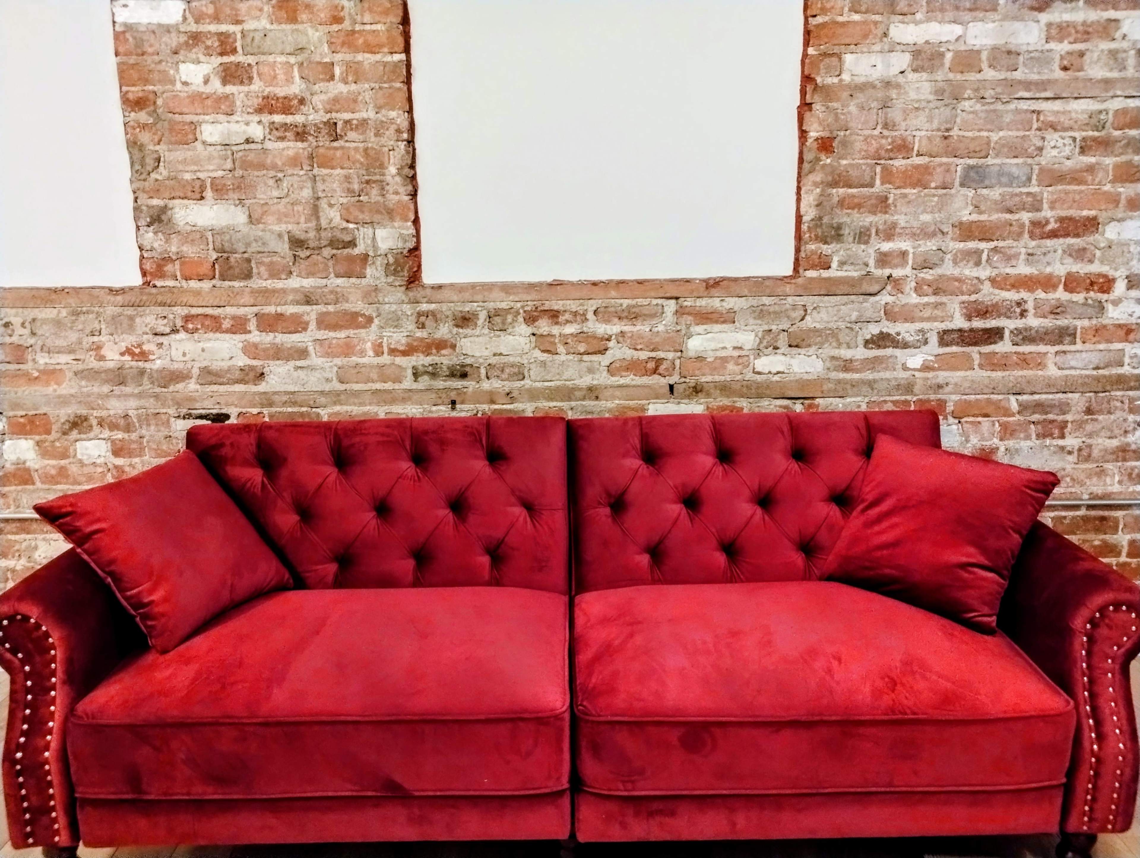 A red tufted sofa is positioned against a backdrop of exposed brick walls with empty frames above it.