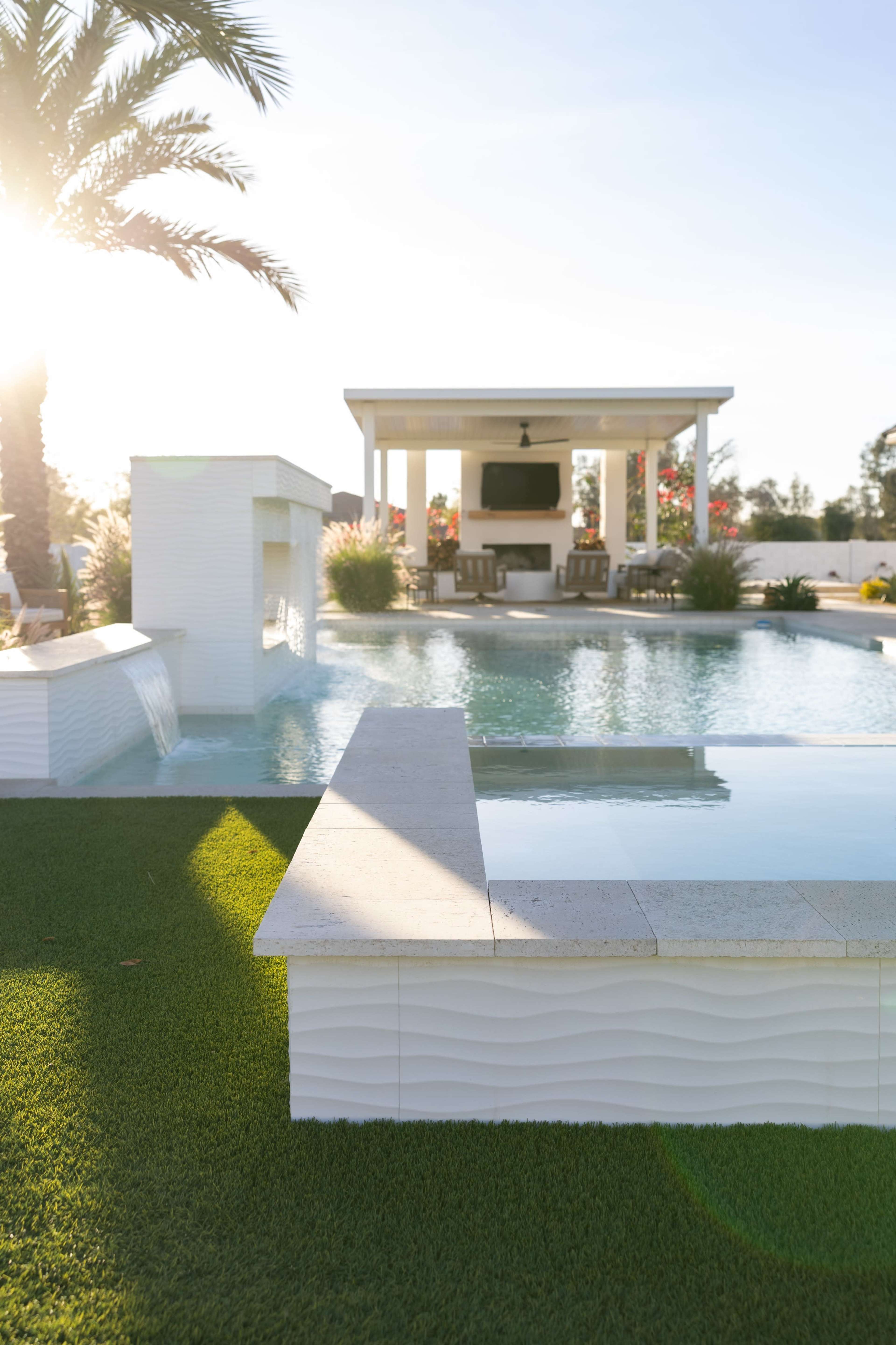 The image shows a modern outdoor pool area with a partially submerged stone edge, a waterfall feature, and a covered seating area surrounded by palm trees and landscaping.