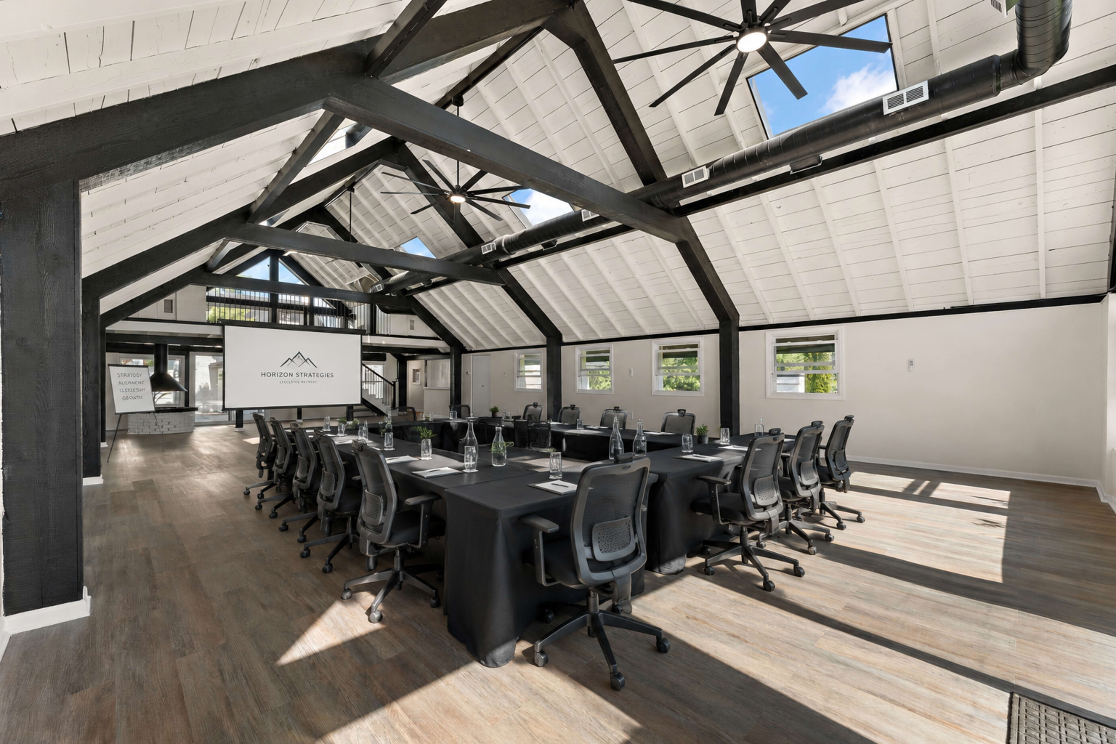A spacious conference room features a long table surrounded by black chairs, with large windows and high ceilings that display exposed beams and ceiling fans.
